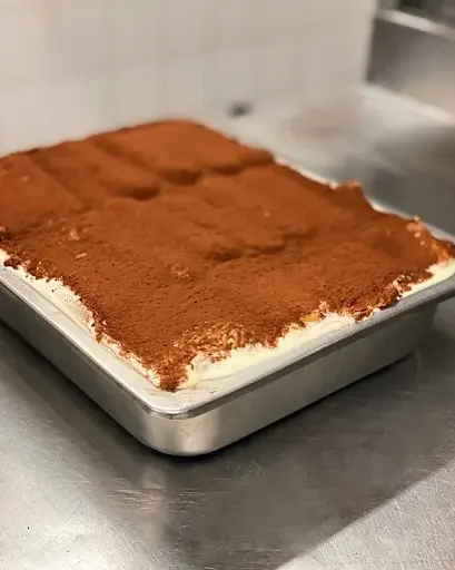 A large metal baking tray filled with freshly made, cocoa-dusted authentic Tiramisu on a kitchen counter, demonstrating the large-batch, made-in-house preparation at Giovanni's Restaurant.