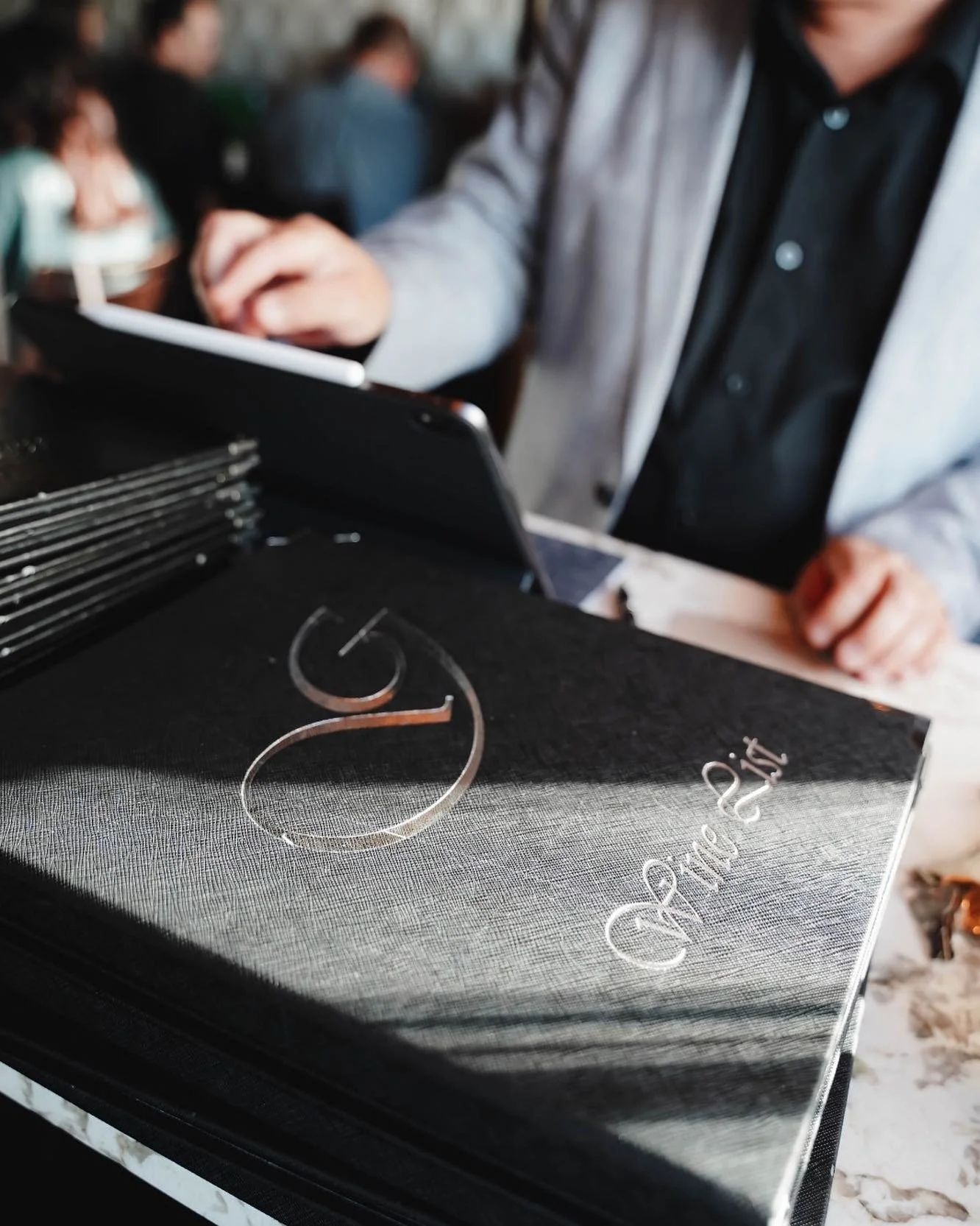 Elegant wine list with embossed “G” logo on a table at an upscale Italian restaurant in Canada, with a guest browsing on a tablet in a refined dining atmosphere.