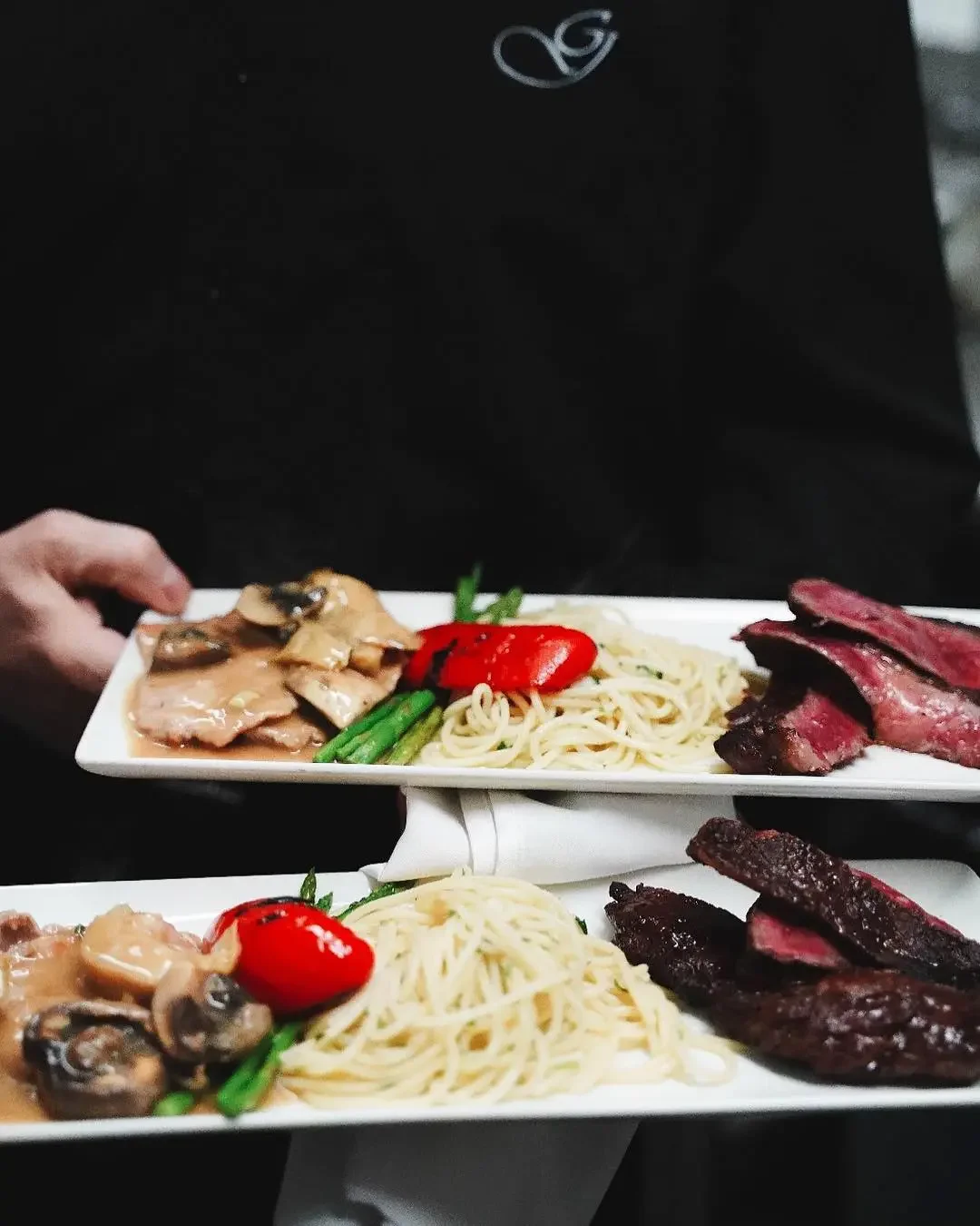 Close-up of a waiter in a formal setting holding two fine-dining plates, featuring a platter of chicken scallopini in mushroom sauce and another with medium-rare steak medallions, both served with angel hair pasta, asparagus, and roasted red pepper.