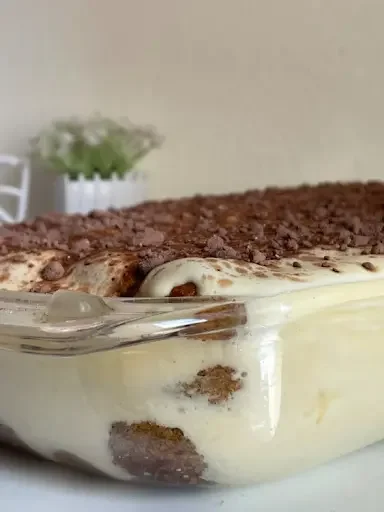 A detailed close-up of layered authentic Italian Tiramisu in a clear glass dish, showing distinct sponge ladyfingers soaked in espresso, thick velvety mascarpone cream layers, and a generous topping of dark cocoa powder and chocolate shavings.