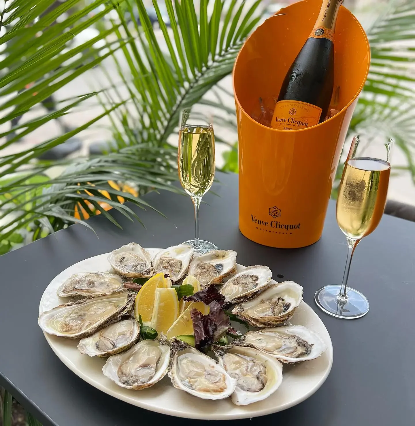 An outdoor patio scene with a large white platter of fresh oysters on the half-shell garnished with lemon, presented alongside two filled champagne flutes and a bottle of Veuve Clicquot champagne chilling in its iconic orange ice bucket.