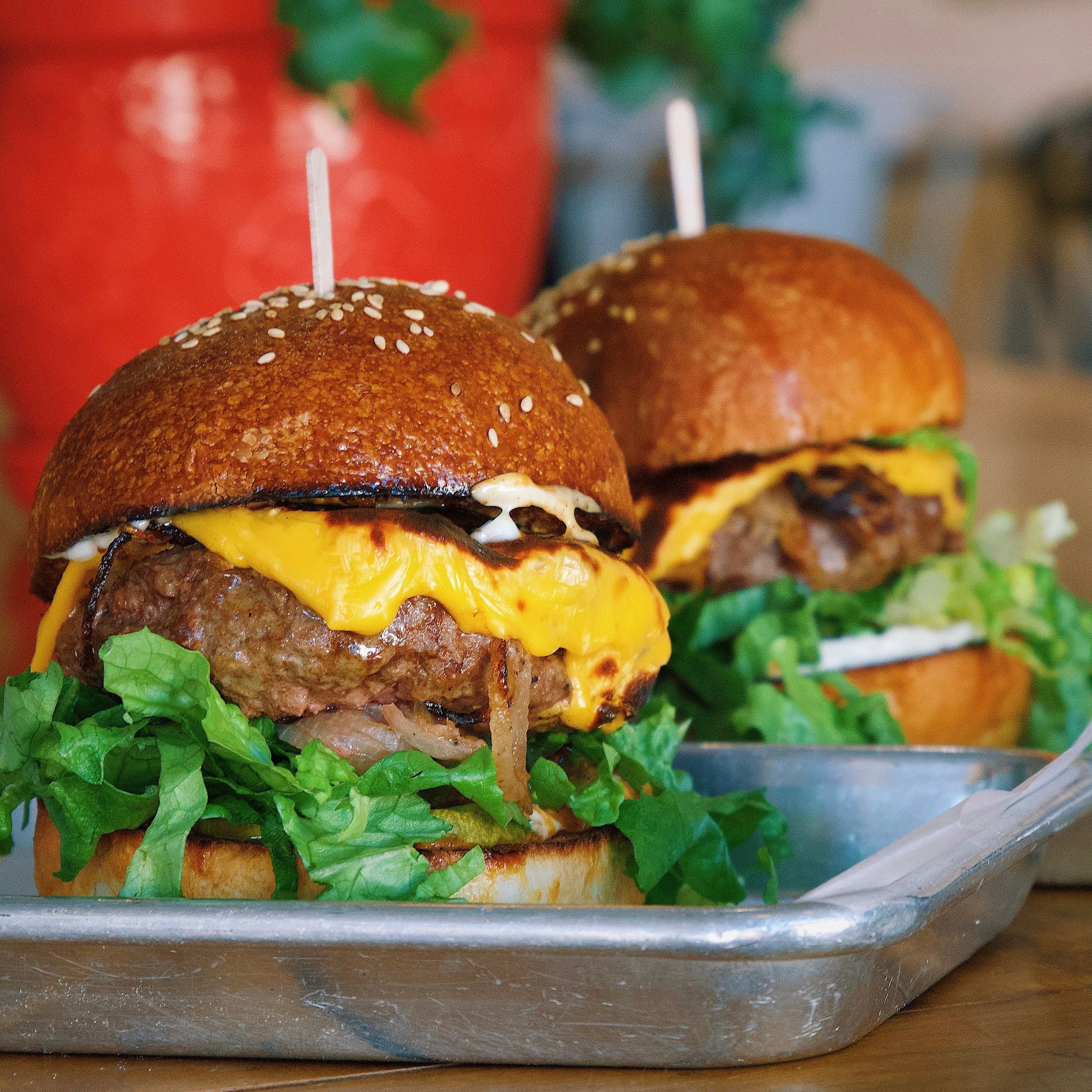 Close-up view of a tray of cheeseburgers with lettuce and toppings, with a blurred background of red and green.