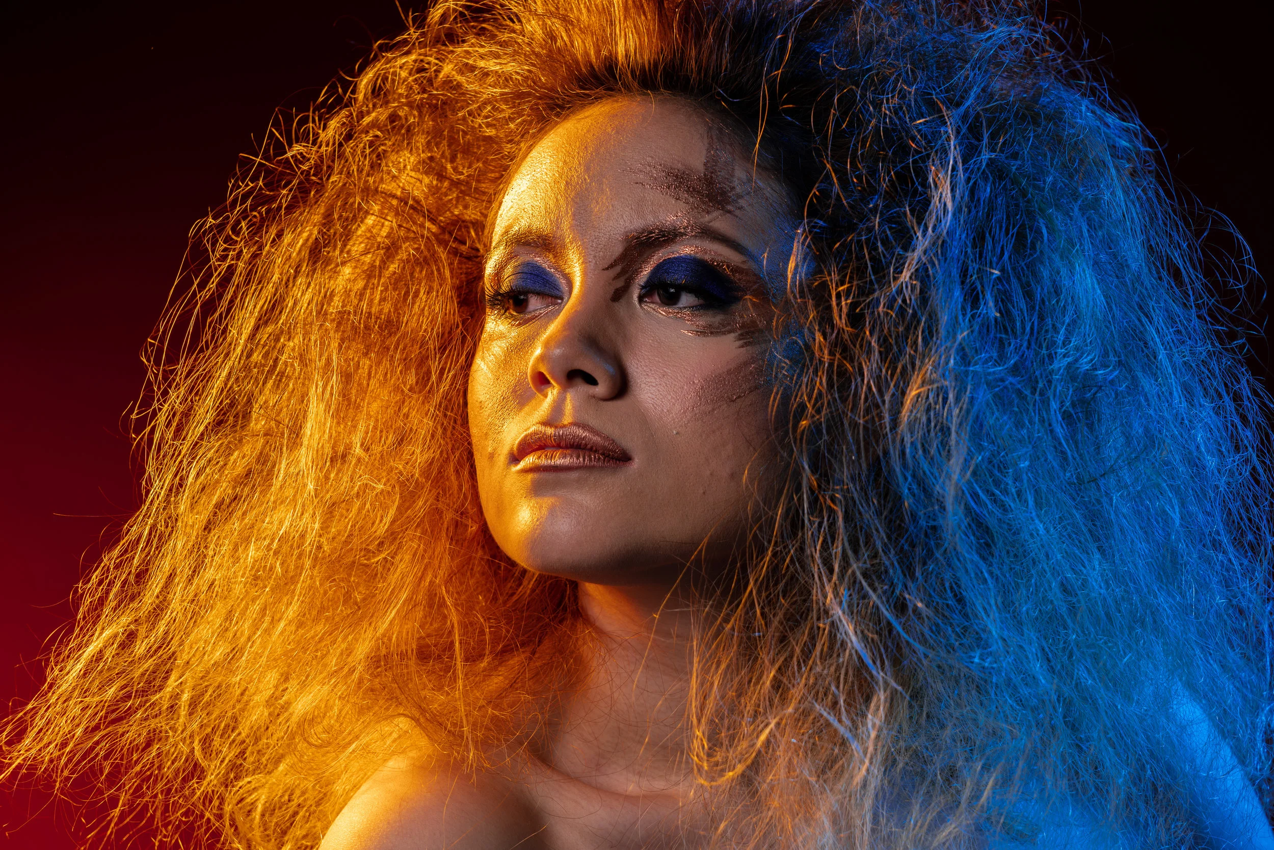 A woman with large, voluminous hair highlighted with blue and orange lighting, wearing bold eye makeup with dark eyeshadow and metallic lipstick.