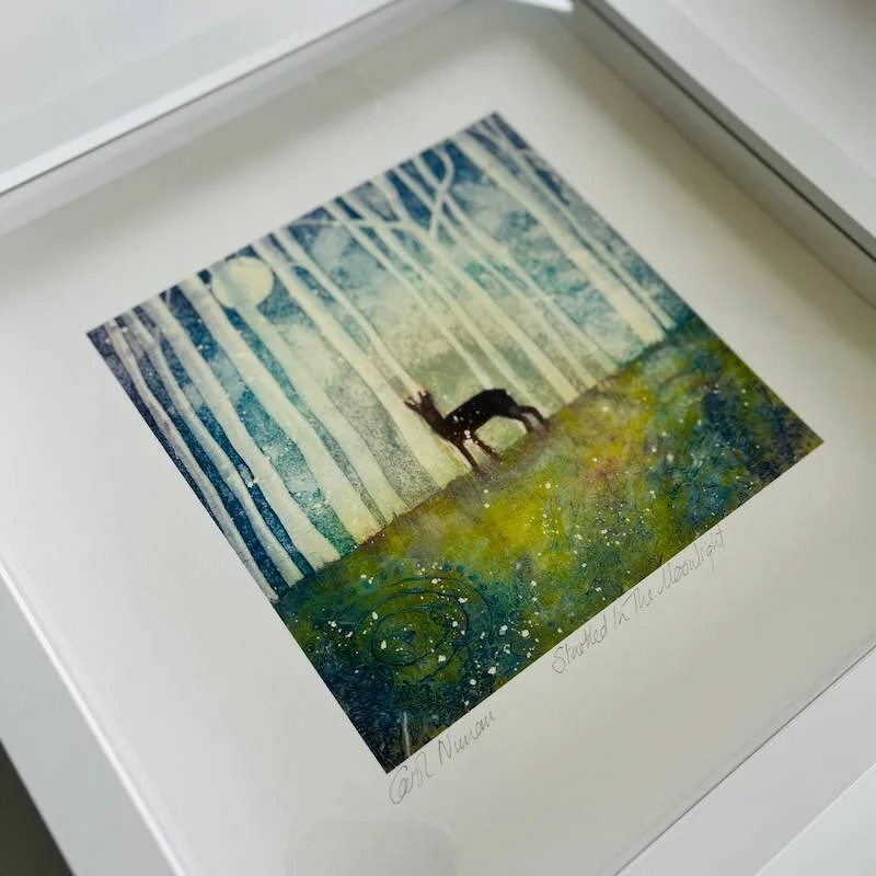 A print of a startled deer in a birch wood with neolithic rock art underfoot in the moonlight in soft shades of blue and green in a white box frame