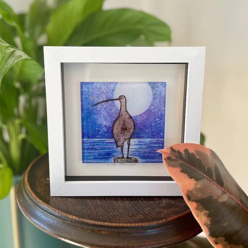 glass-art-tile-carol-nunan-in-the-moonlightglass art tile - on the hunt - a collagraph print of a curlew under a full moon looking out across the sea framed in a white box frame on a shelf
