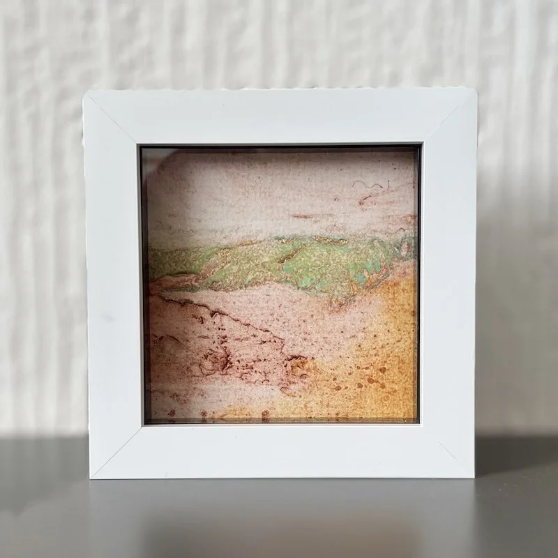 A box framed semi-abstract collagraph mini print using textured wall papers to create rocky strata of a hillside on the horizon