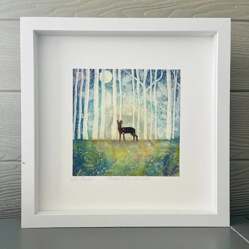 A print of a startled deer in a birch wood with neolithic rock art underfoot in the moonlight in soft shades of blue and green in a white box frame
