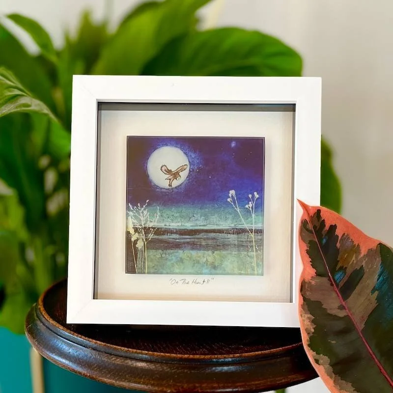 glass art tile - on the hunt - a collagraph print featuring an owl hovering up over a moorland landscape under a full moonframed in a white box frame on a shelf