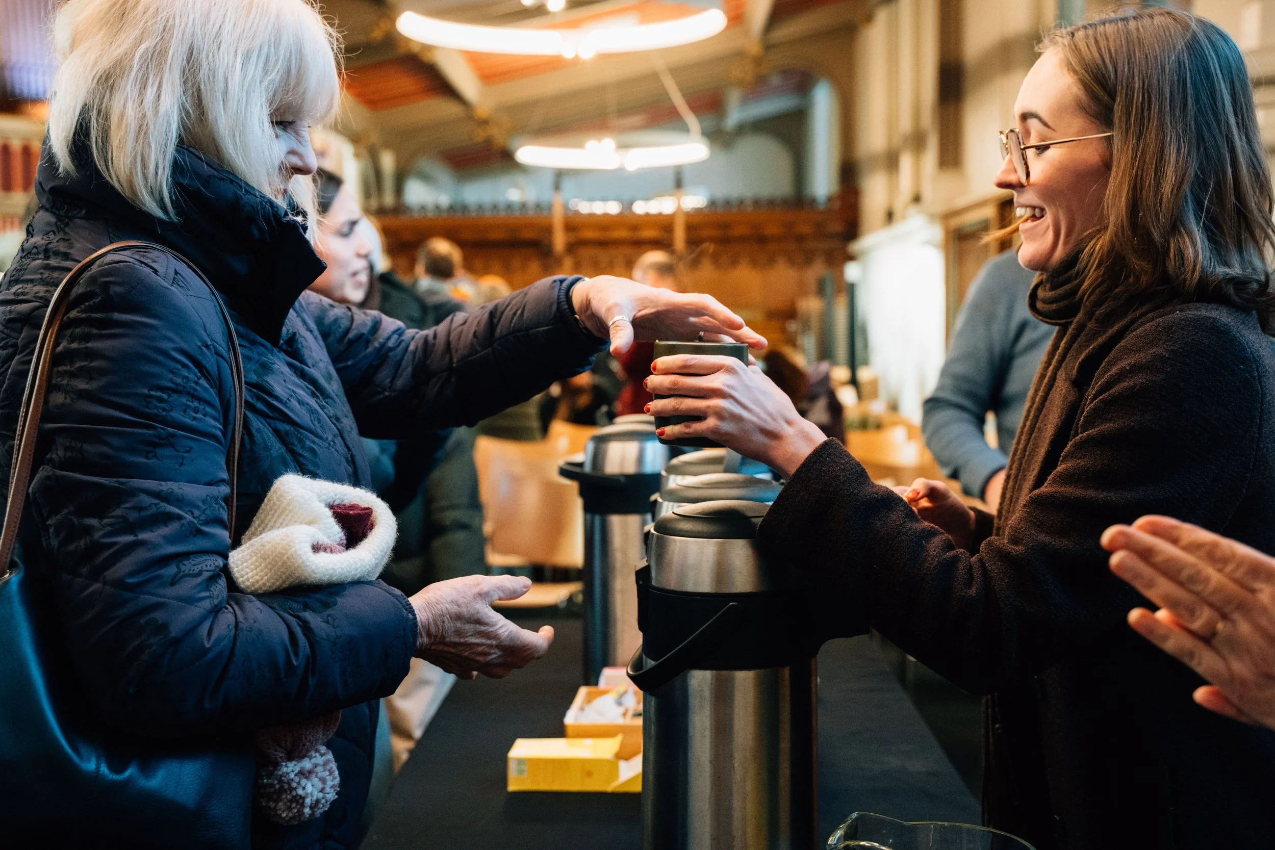 A woman in a black jacket receiving a hot beverage from a smiling woman behind a counter at a busy indoor event or cafe.