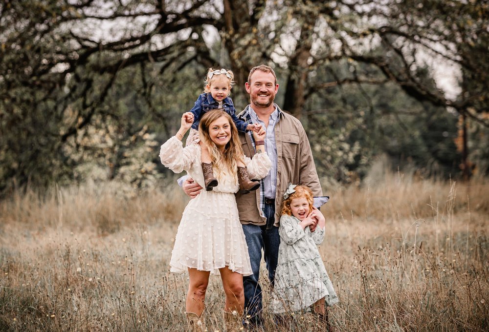 A family of four in a golden field near Sandy, Oregon. The dad carries a little girl on his shoulders while the mom laughs beside him and an older girl giggles in front.