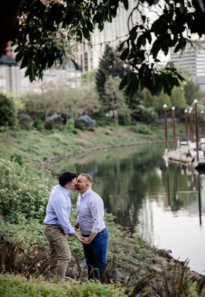Two grooms kiss along the Willamette River with the downtown Portland skyline rising behind them