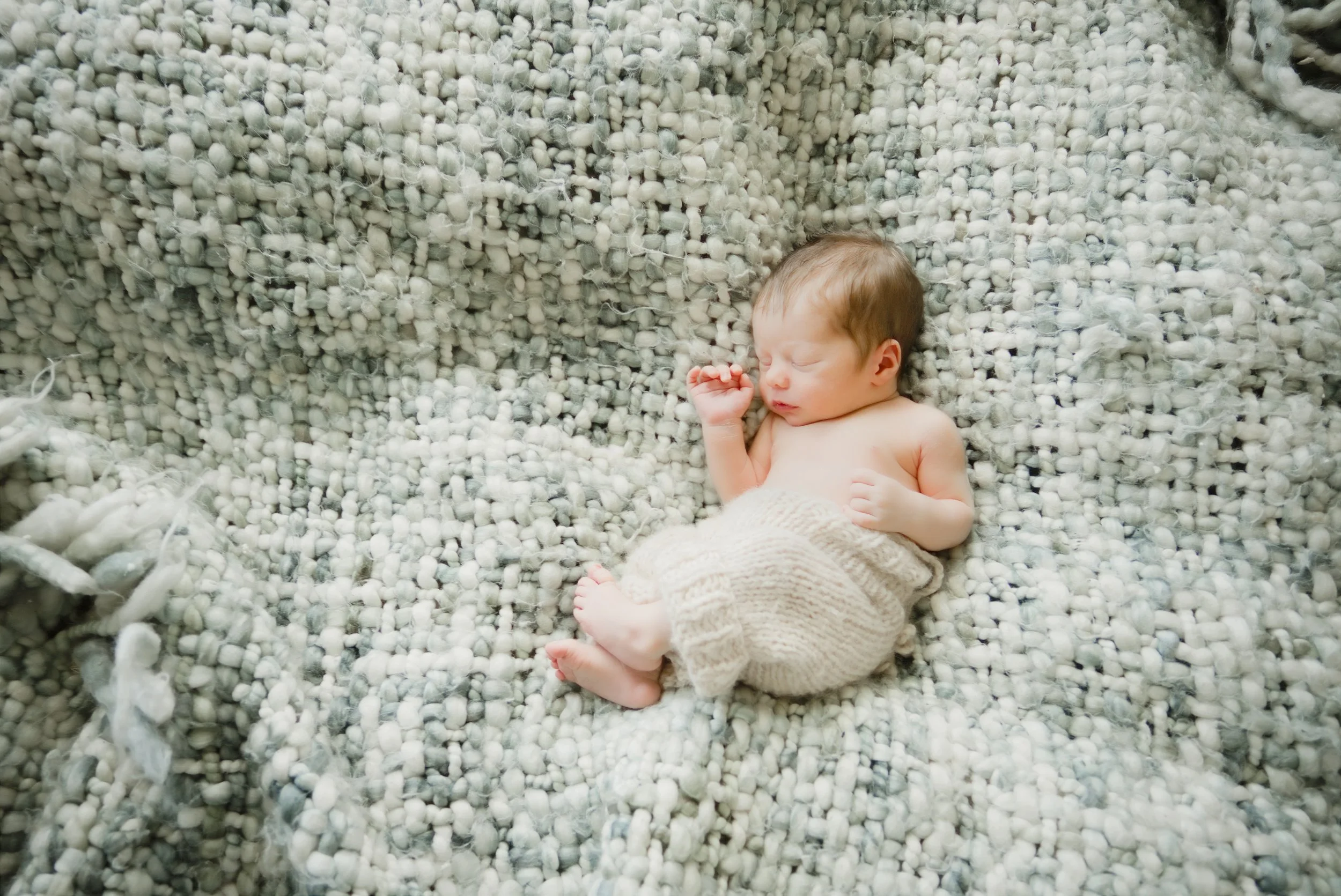 Newborn curled up in knit pants on a textured cream blanket