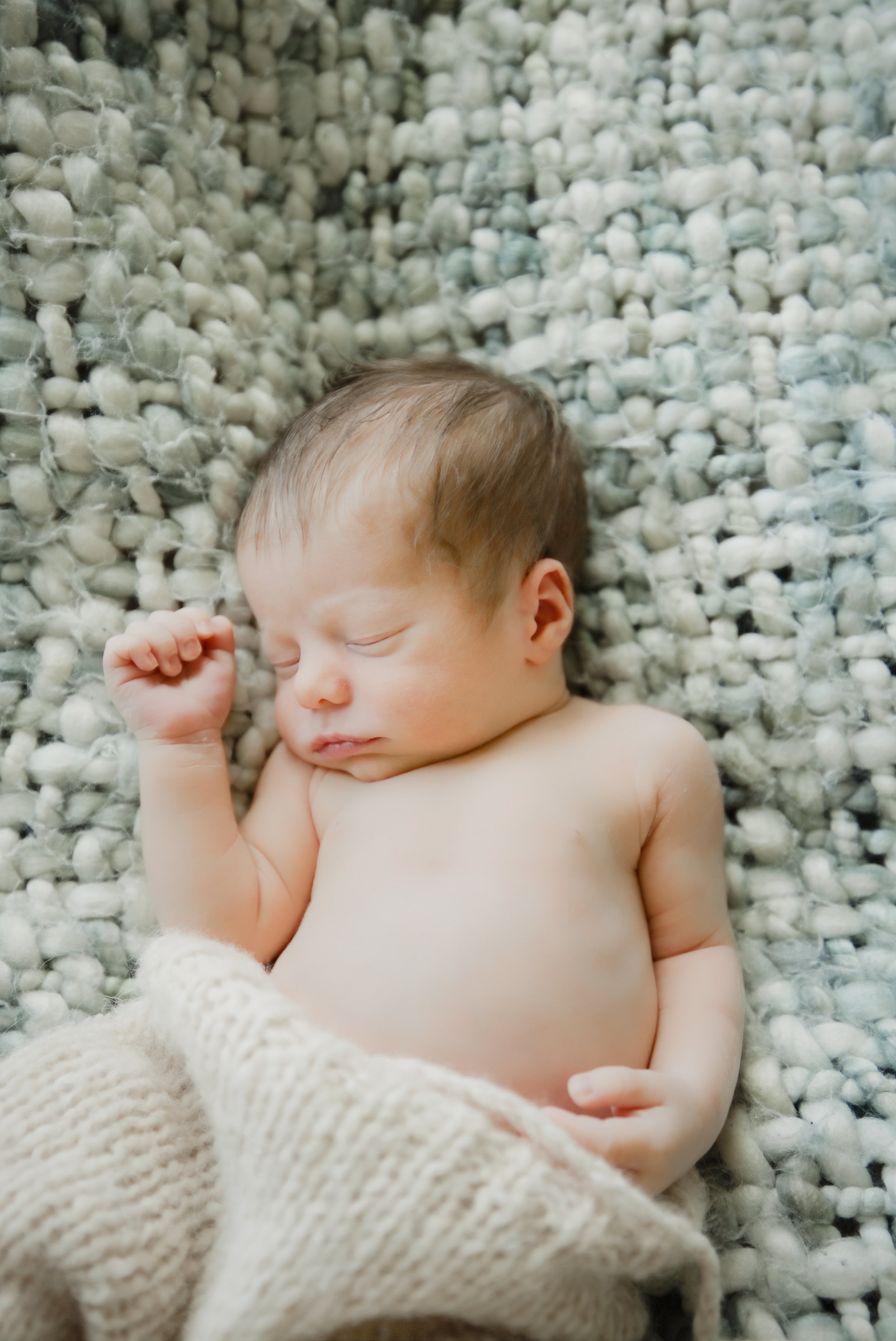 One week old baby sleeps on a chunky blue grey blanket with one arm stretched overhead