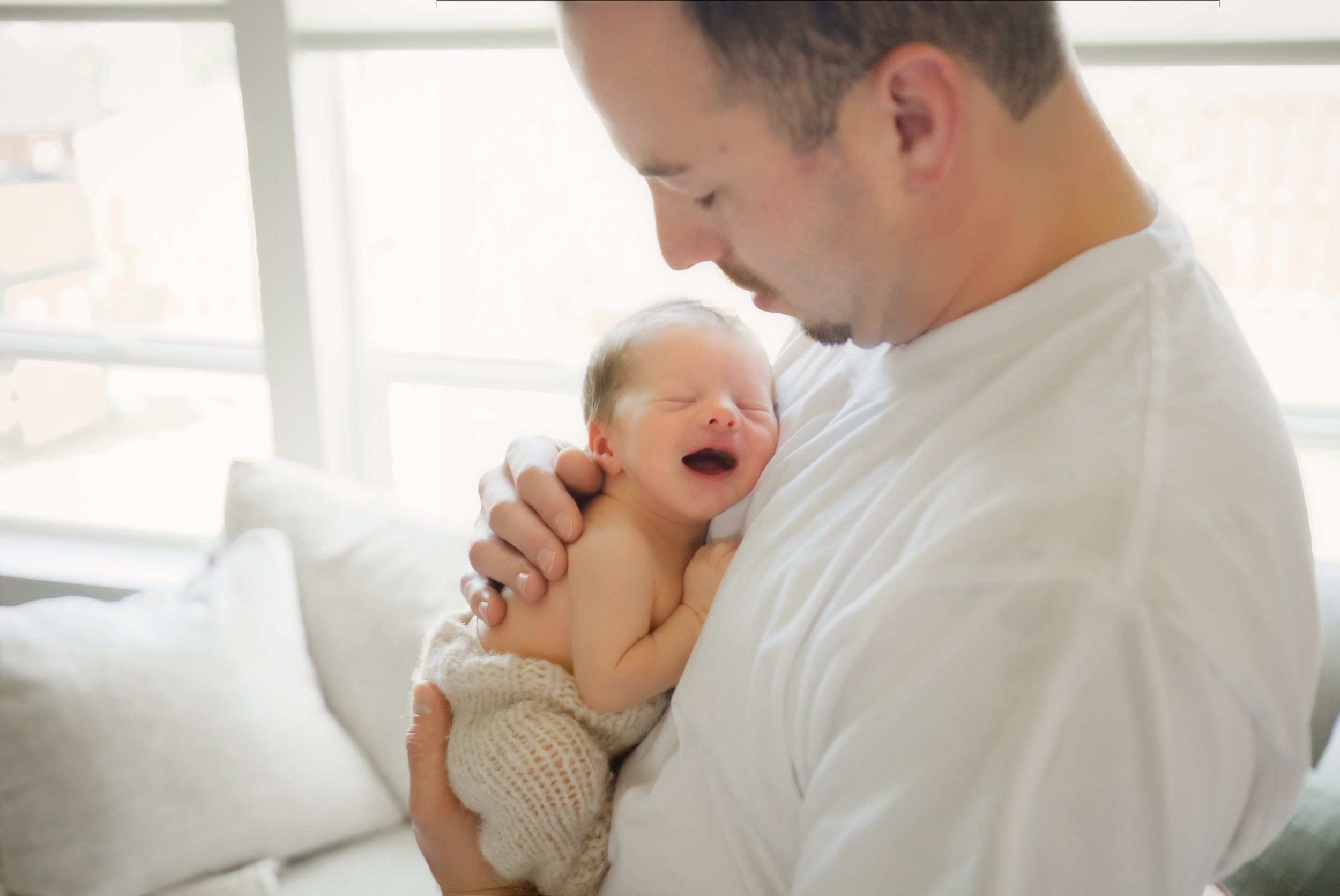 Dad soothes his newborn son by a bright window during an in home newborn session