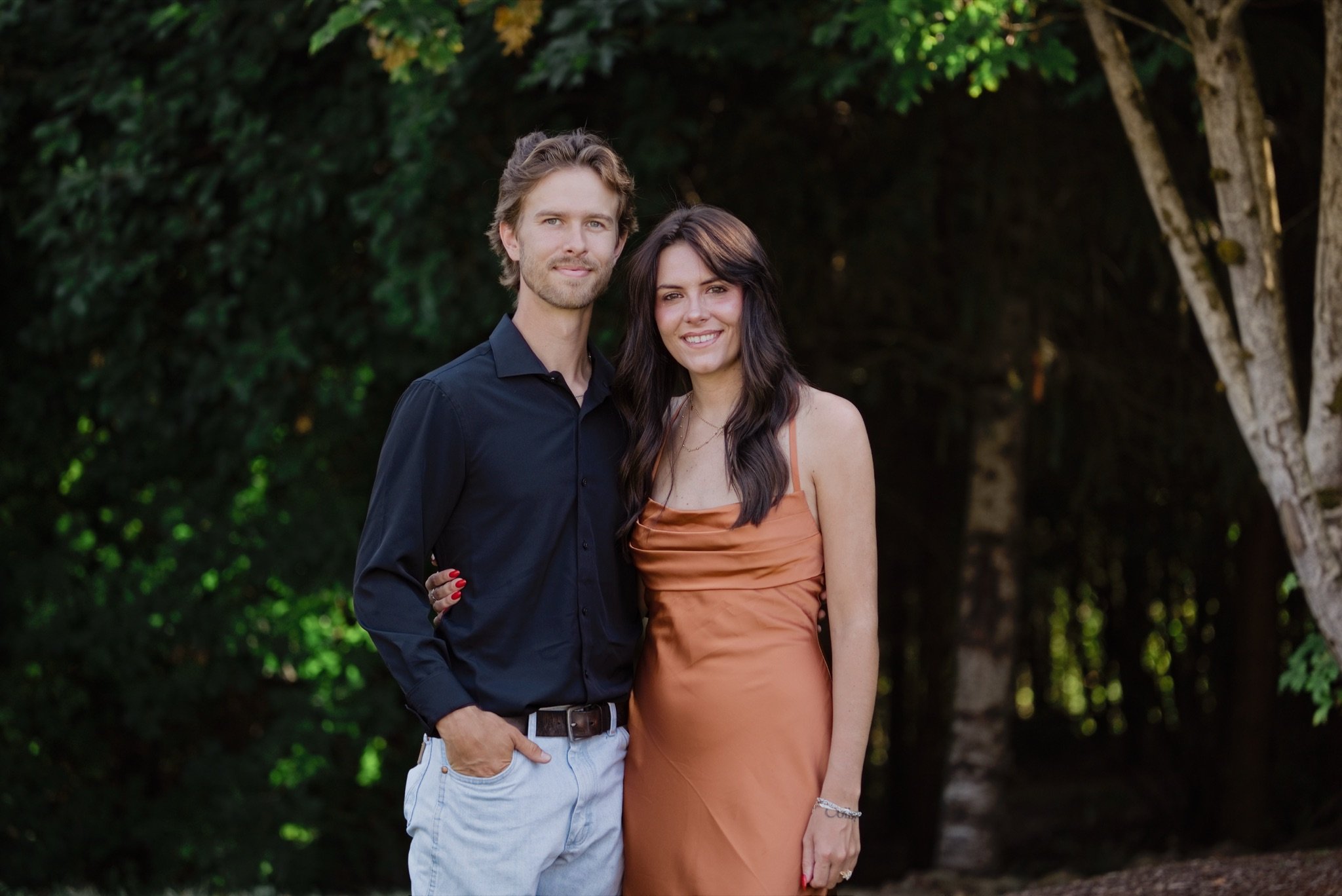 A couple from the wedding party in black and rust under the trees