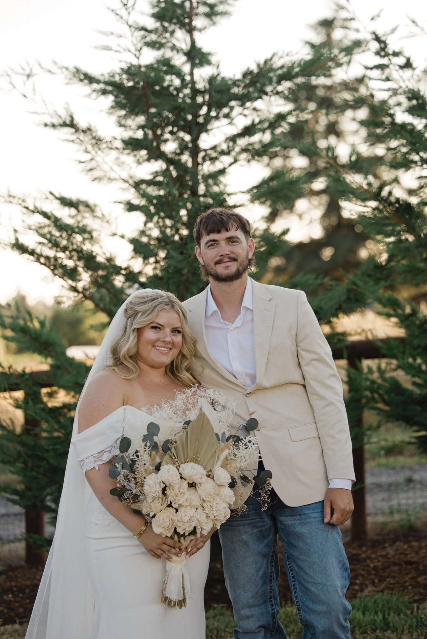 Sarah and Reed smiling in front of the firs with her bouquet