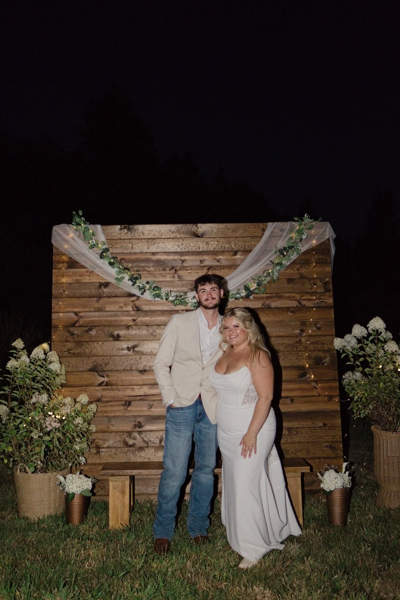 Sarah and Reed in front of the wooden backdrop with greenery at night