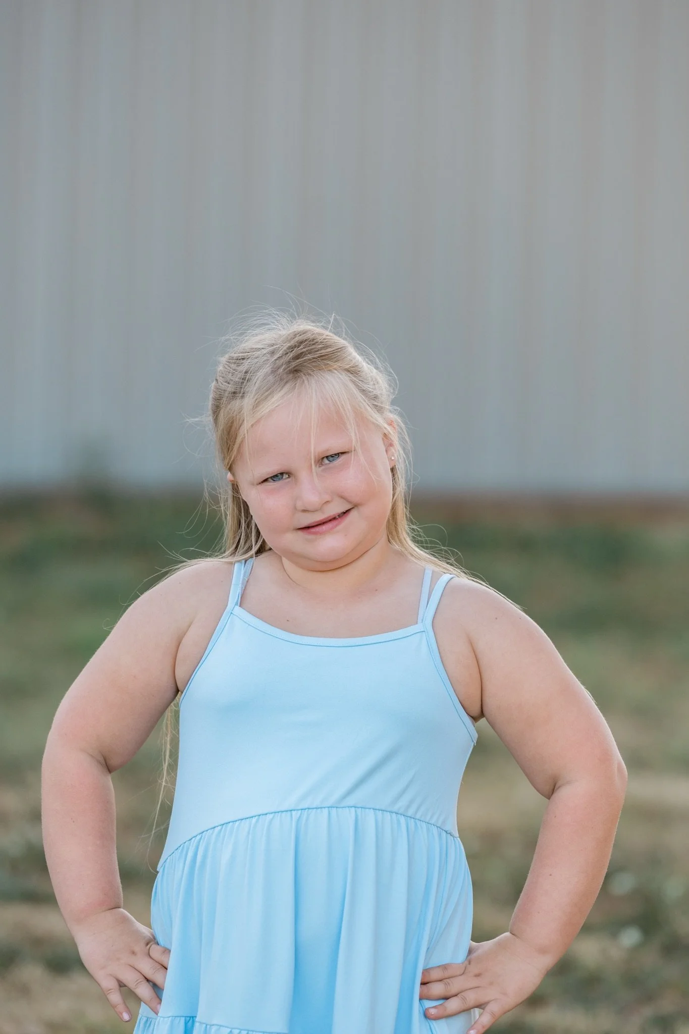 A little girl in a light blue dress with her hands on her hips in the field