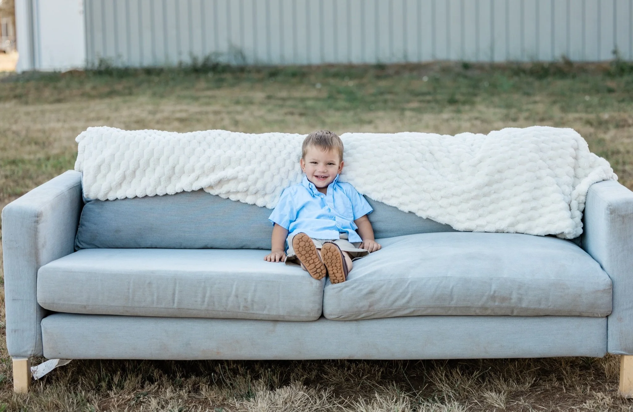 A toddler in blue sitting alone on the big gray couch in the field