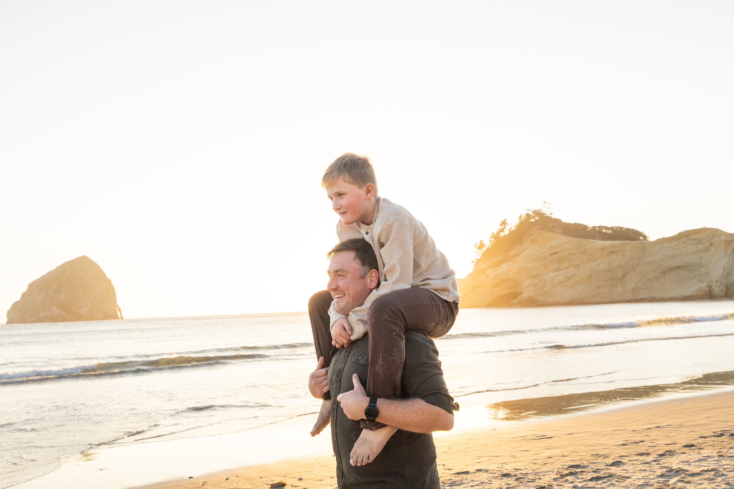 Boy laughs on his dad's shoulders at the surf line with Haystack Rock and the cape behind them