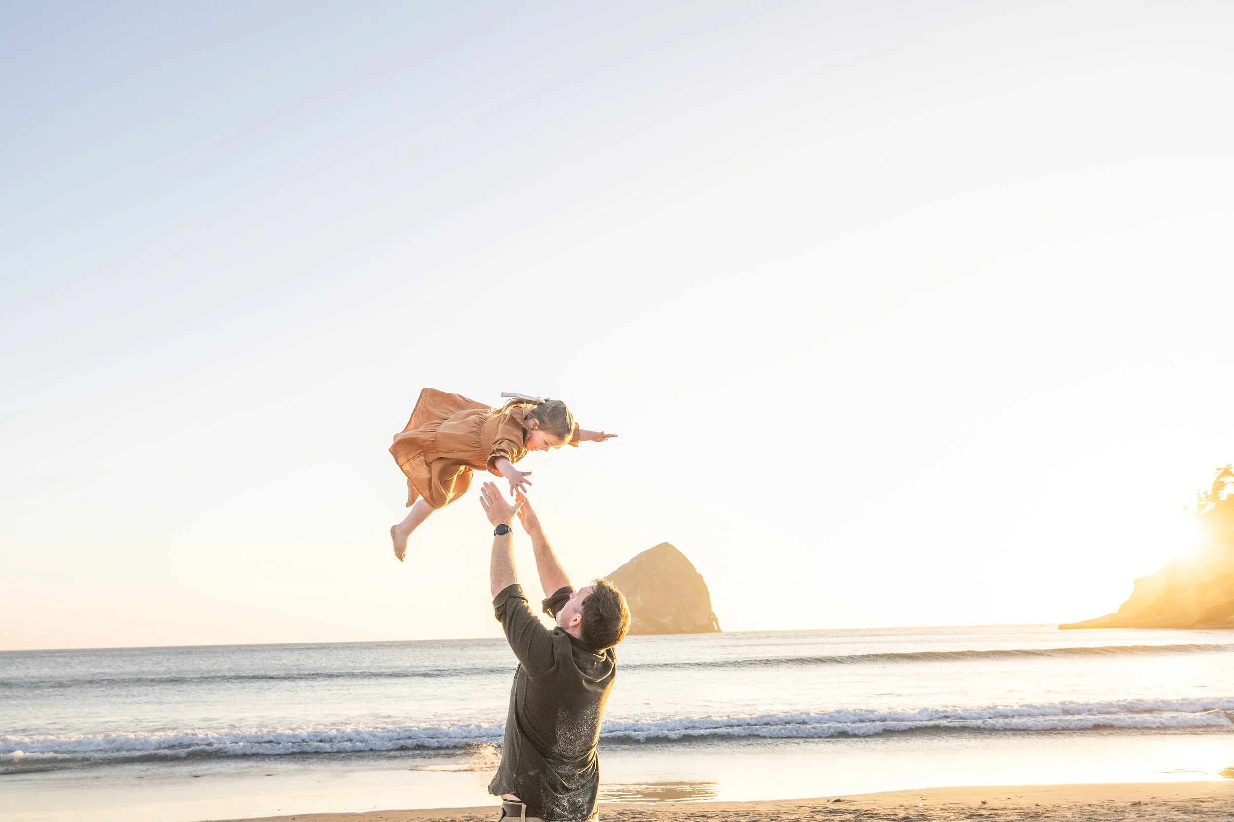 Dad tosses his toddler into the air at the tide line with Haystack Rock behind her at sunset