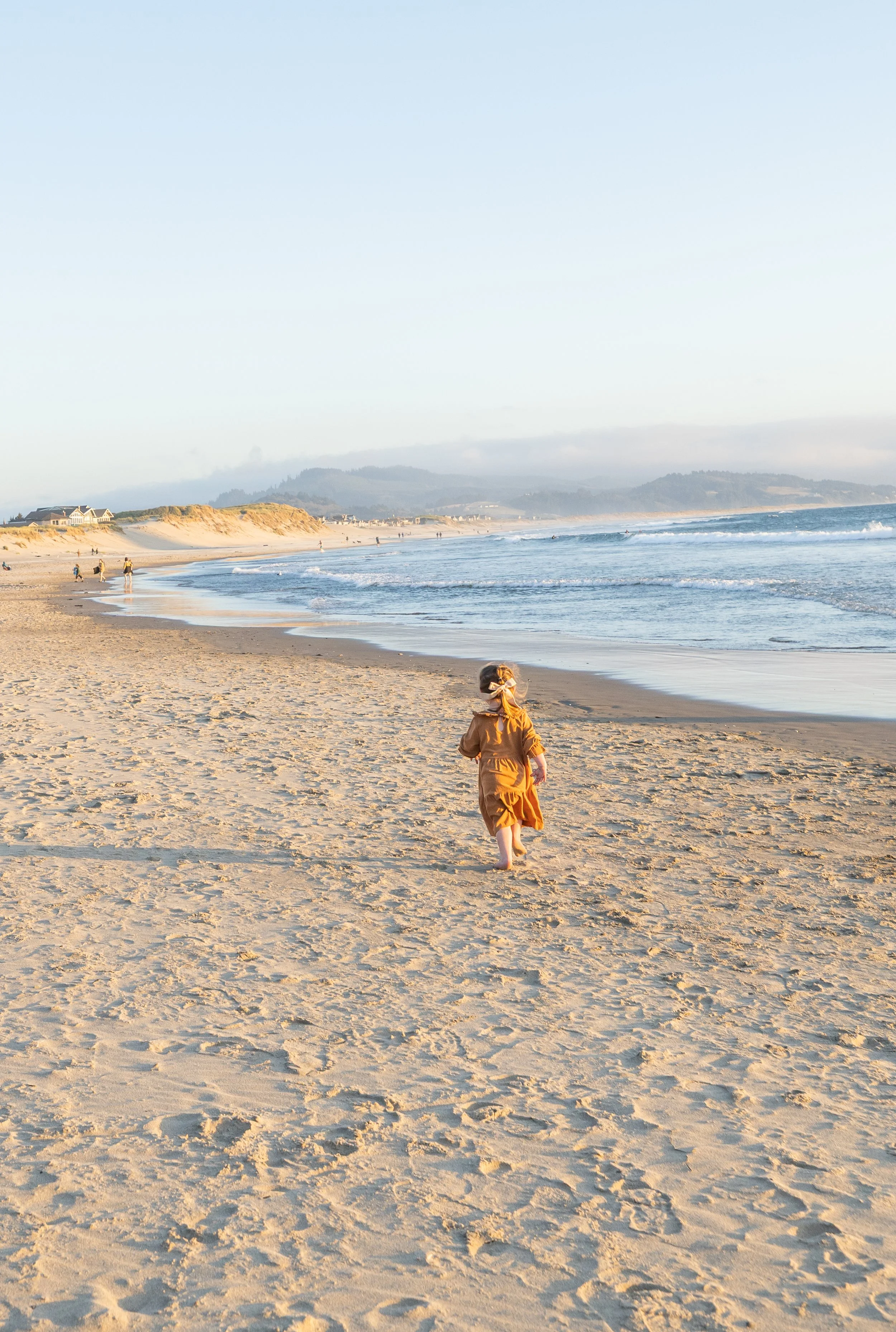 Toddler in a rust dress runs down the beach toward the camera at golden hour in Pacific City