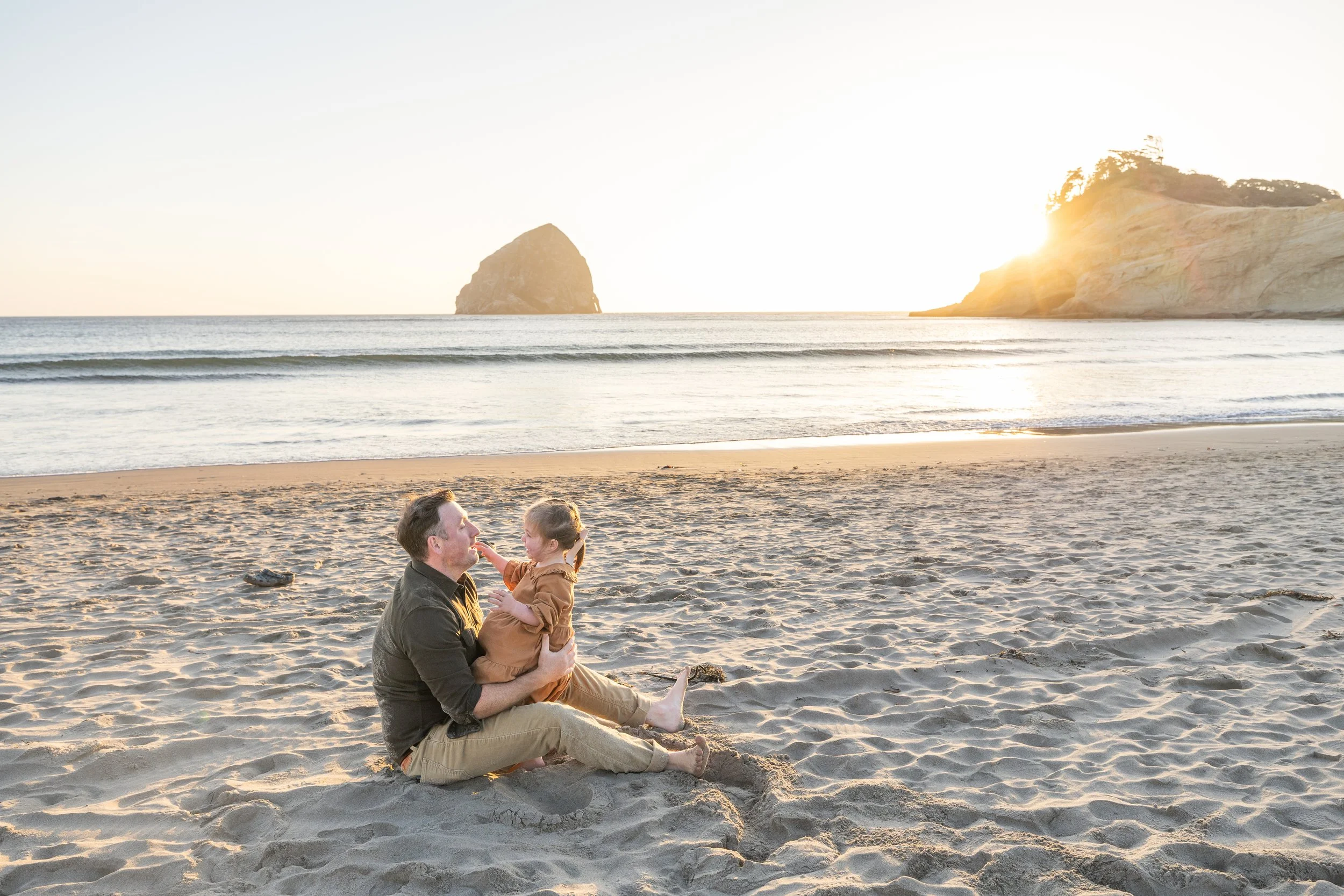 Dad sits on the beach holding his daughter nose to nose with Haystack Rock in the distance