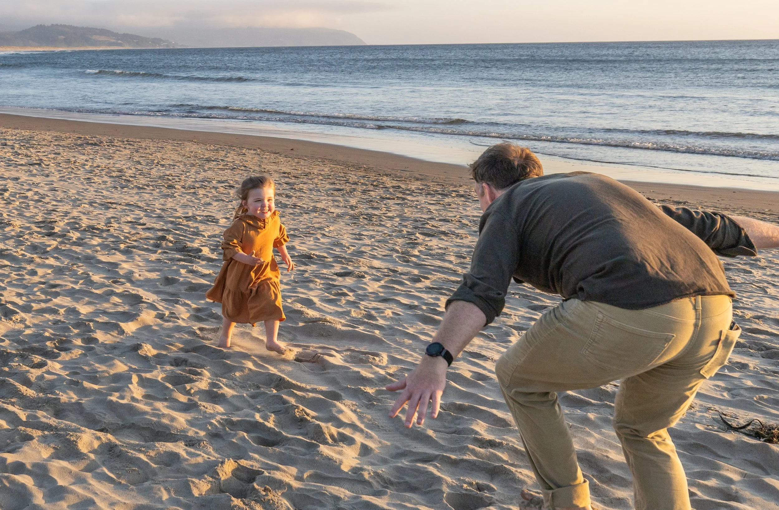 Dad crouches with open arms as his toddler runs to him across the sand at Cape Kiwanda