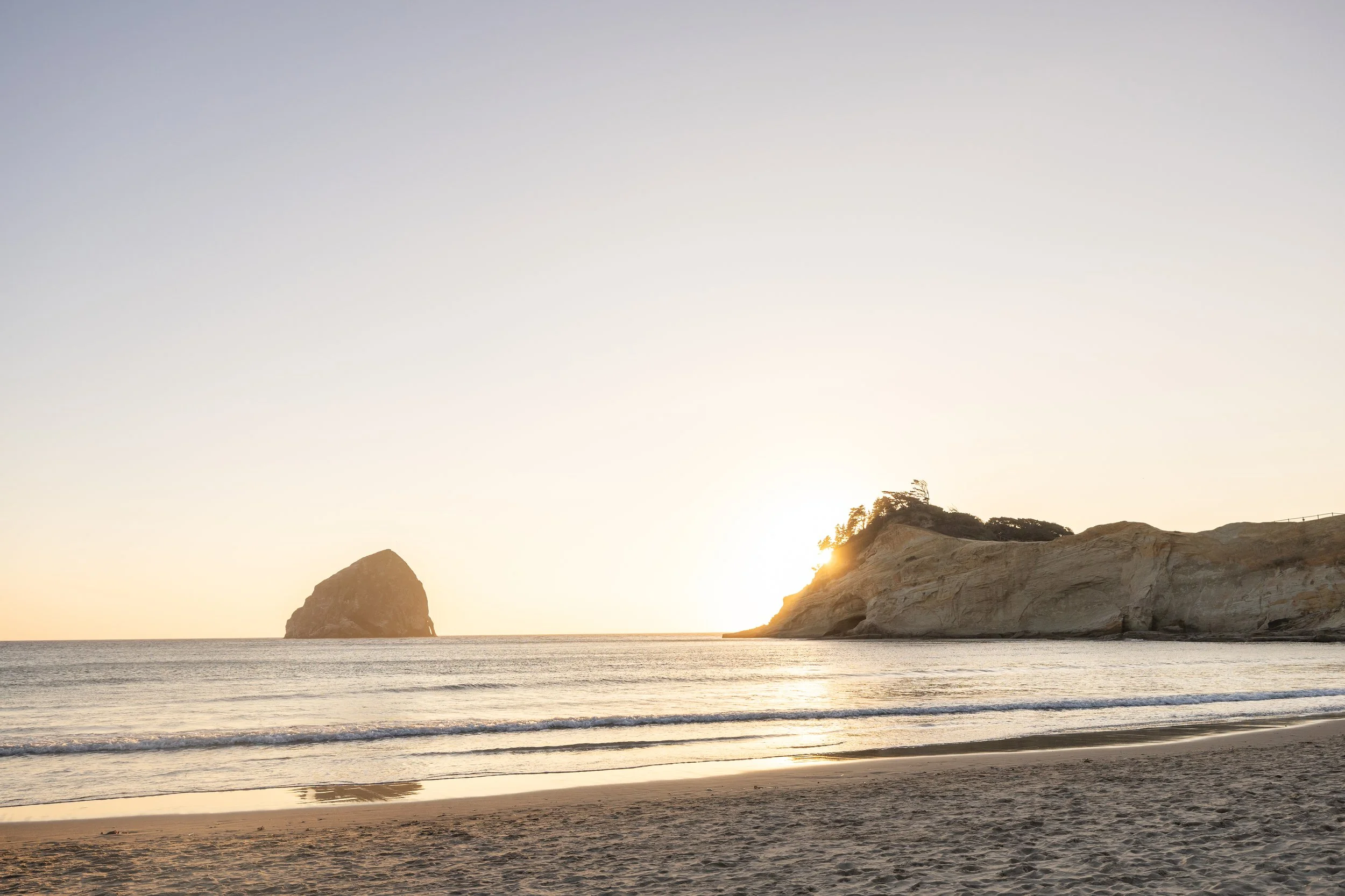Haystack Rock and the Cape Kiwanda headland glow at sunset over calm waves in Pacific City, Oregon