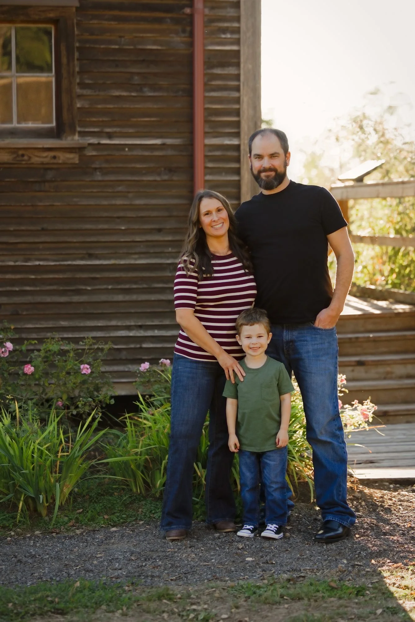 Family of three beside the barn with pink roses still blooming along the garden fence