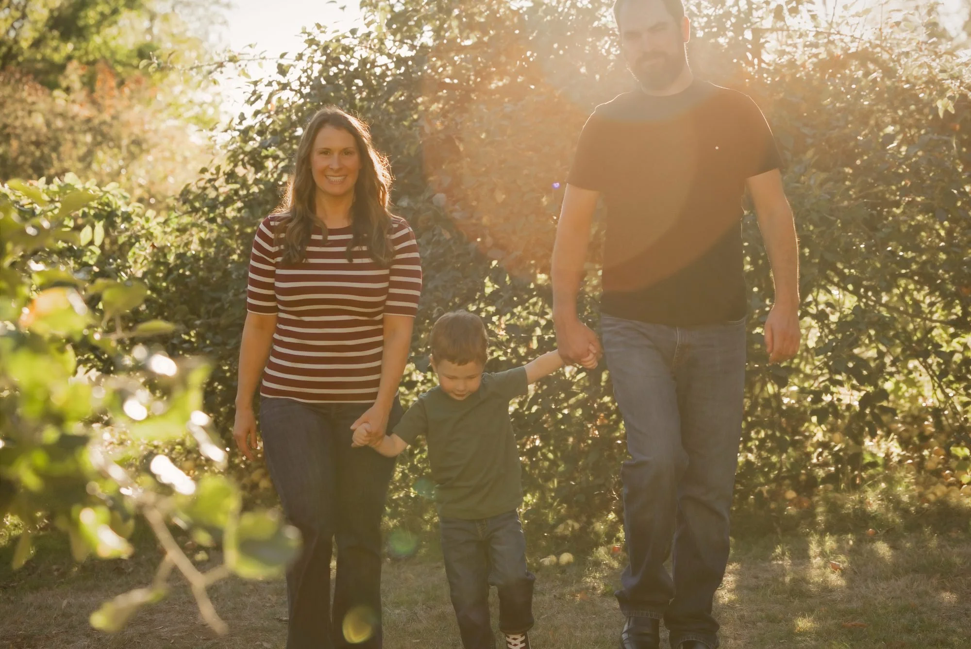 Parents holding their baby boy in front of the white farmhouse with backlit trees at sunset