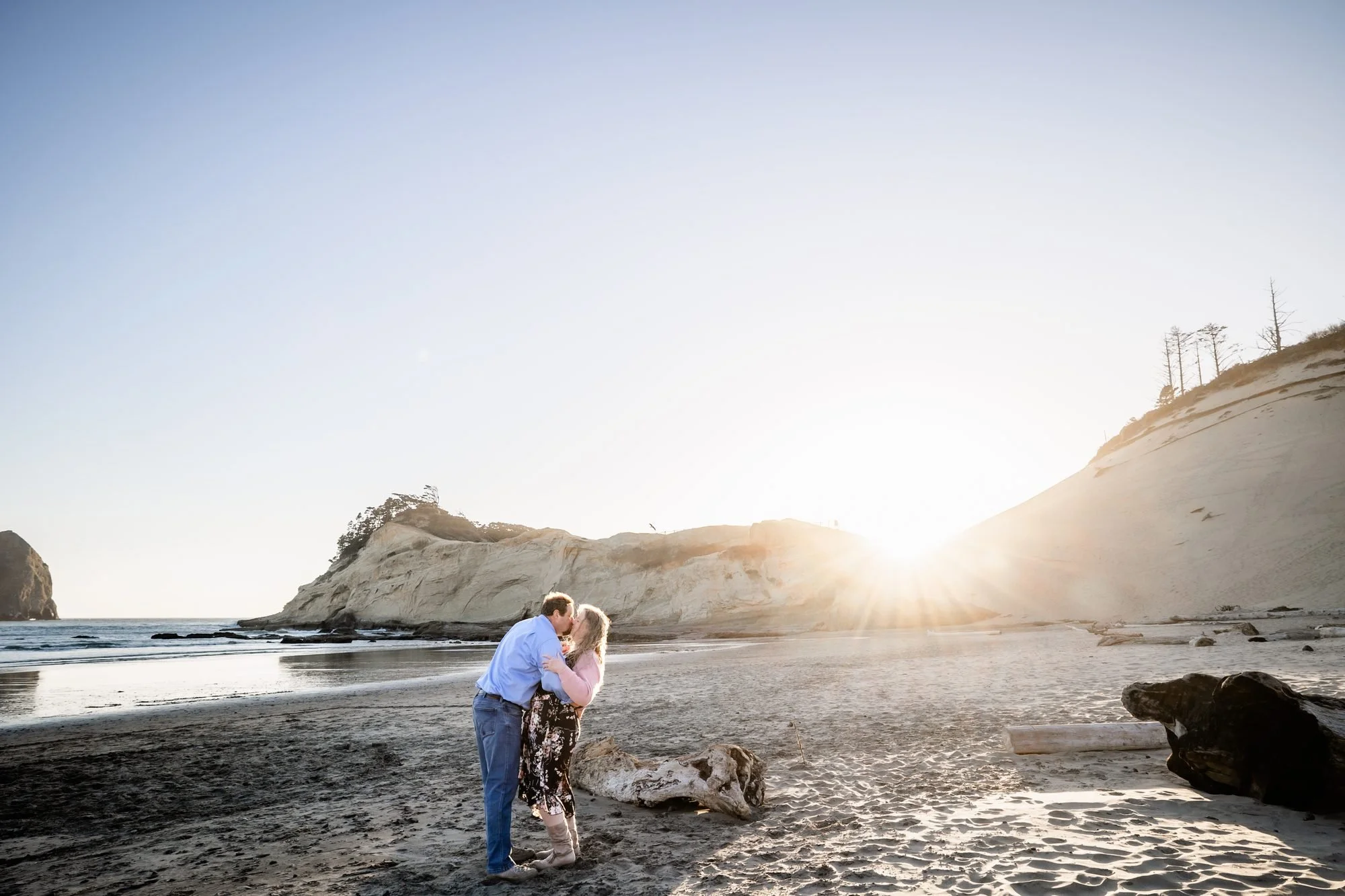 The couple embracing on the beach with the sun setting behind the Cape Kiwanda headland