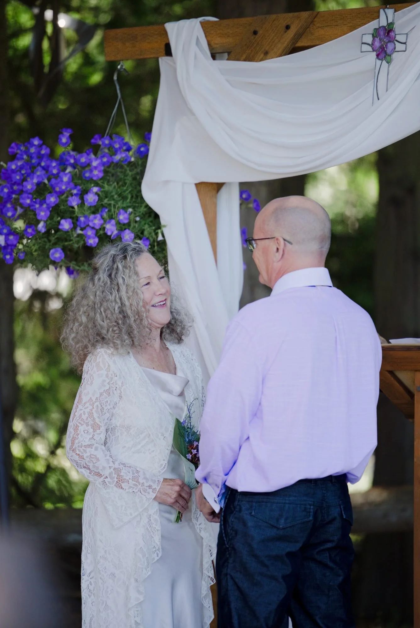 Trudy and Michael holding hands under the draped wooden arch with purple petunias