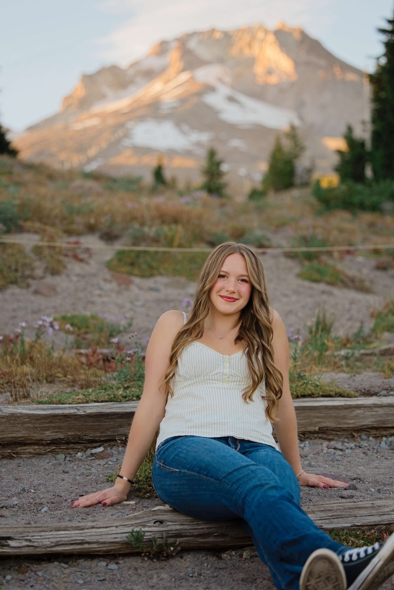 This senior sitting on a log in a white tank top and jeans below Mt. Hood