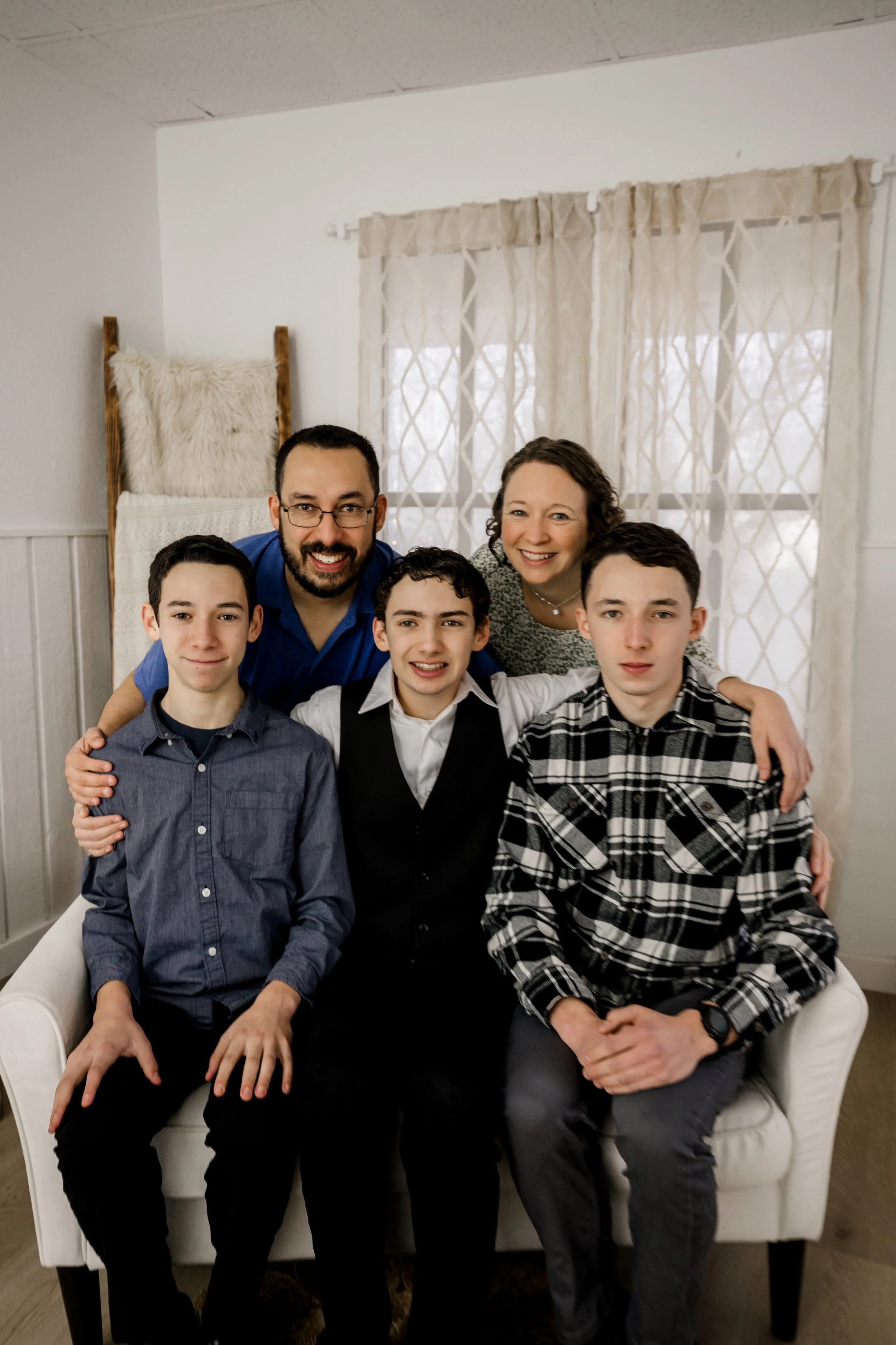 A family of five posing together on a white sofa in a room with white walls and beige curtains, smiling at the camera.