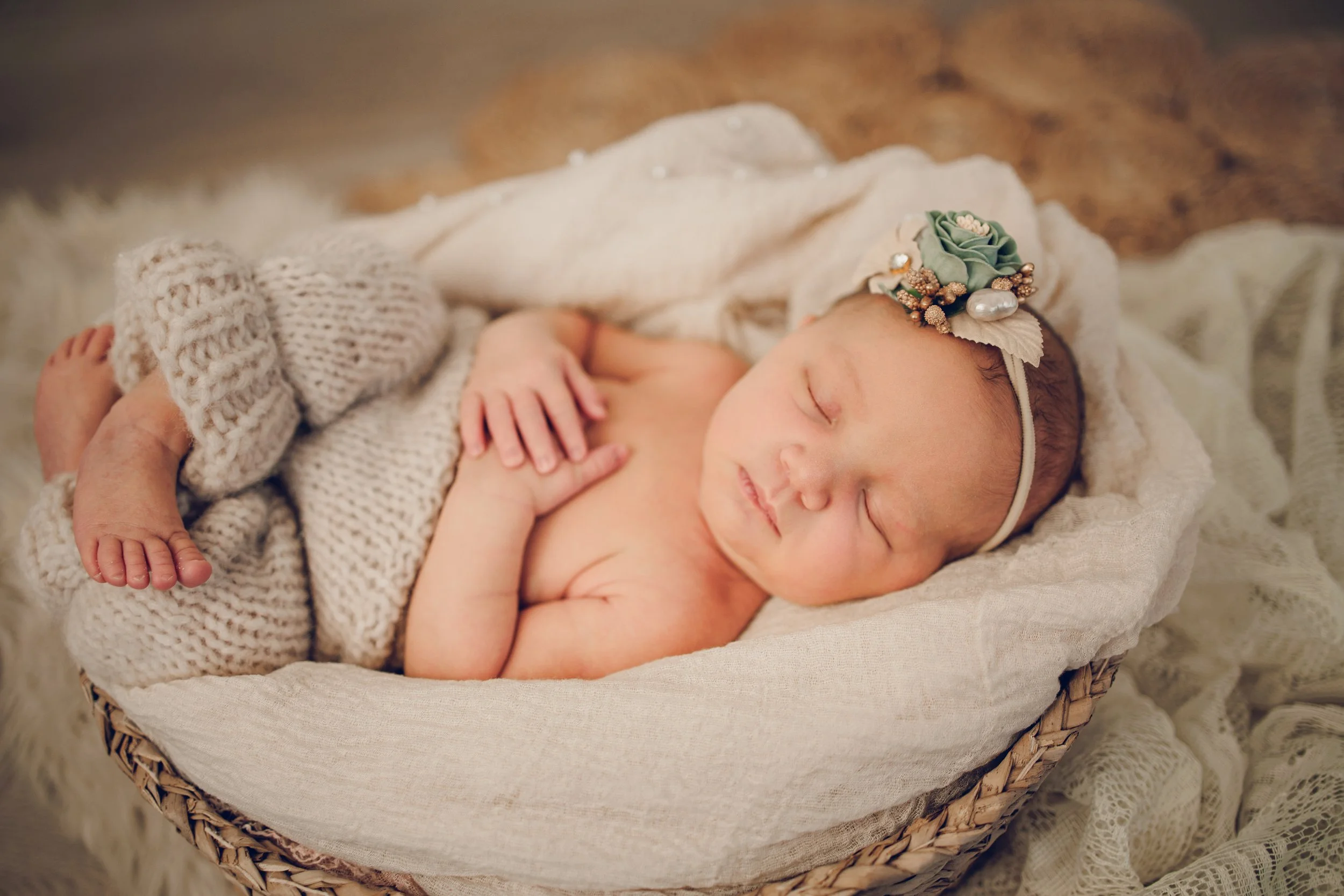 A sleeping newborn baby girl lying in a woven basket with a white blanket, wearing a floral headband with green and pearl accents.