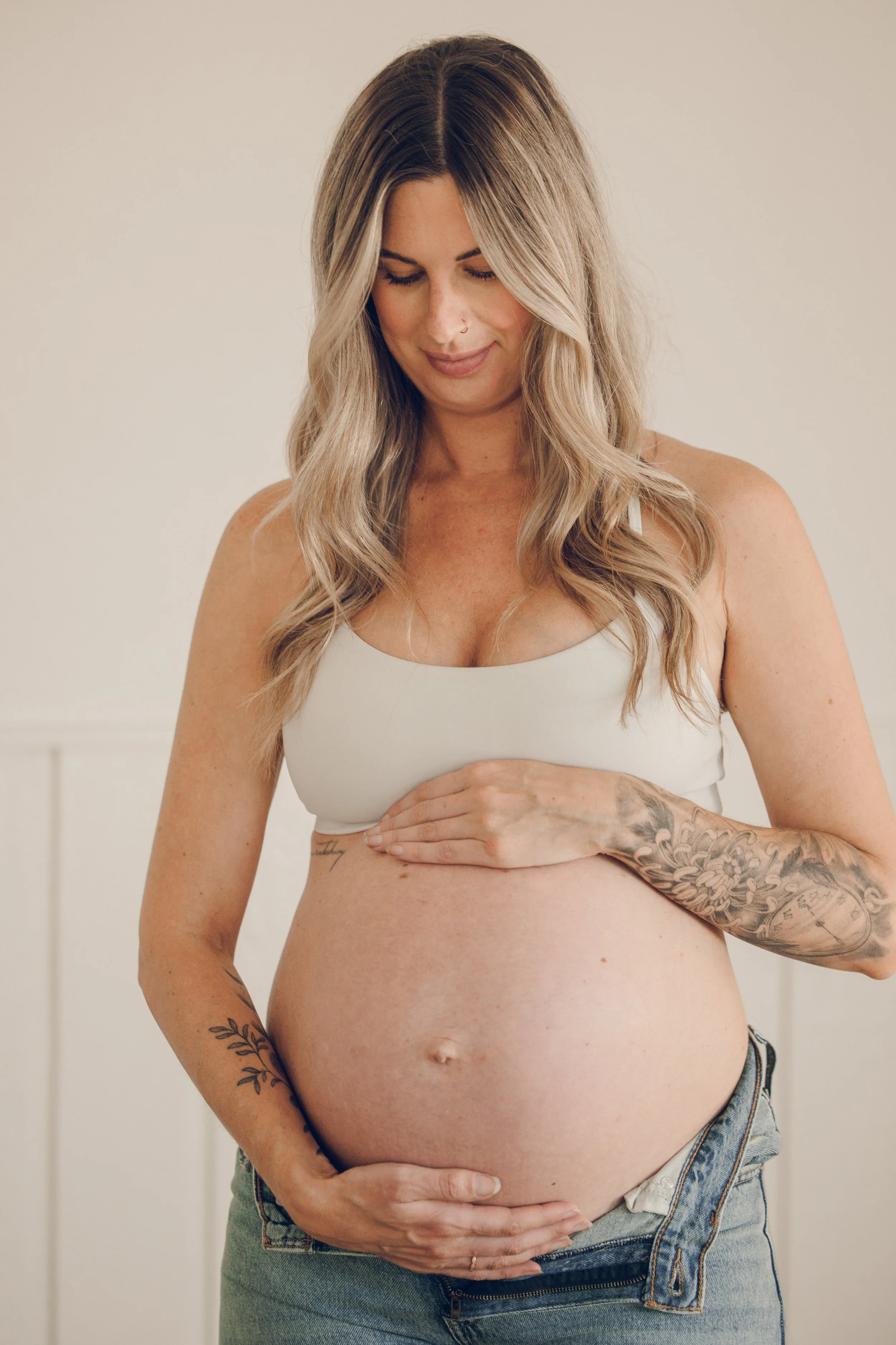 A pregnant woman with long wavy blonde hair, tattoos on her arms, wearing a white sports bra and jeans, standing indoors with a neutral background, looking down at her belly and gently holding it.