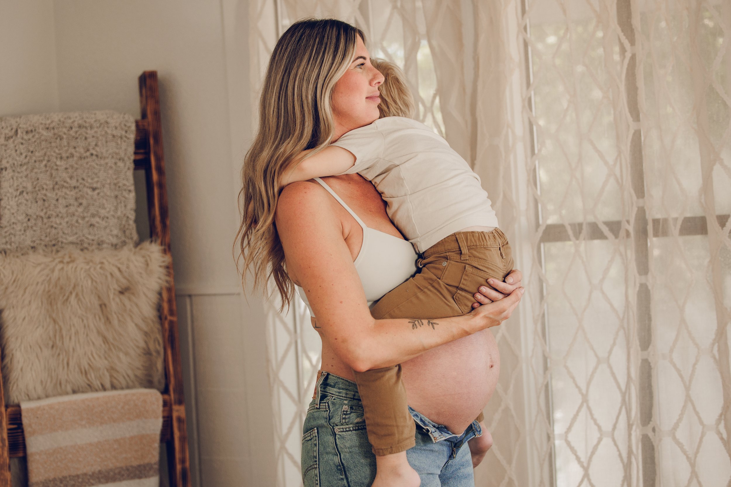 A pregnant woman holding and hugging a young child by a window with lace curtains, with warm natural lighting.
