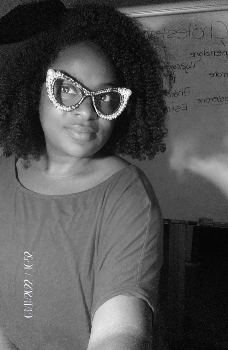 A woman with curly hair wearing glasses with rhinestones, smiling softly in a black-and-white photo. She is wearing a short-sleeved shirt and is standing near a whiteboard with handwritten notes in the background.
