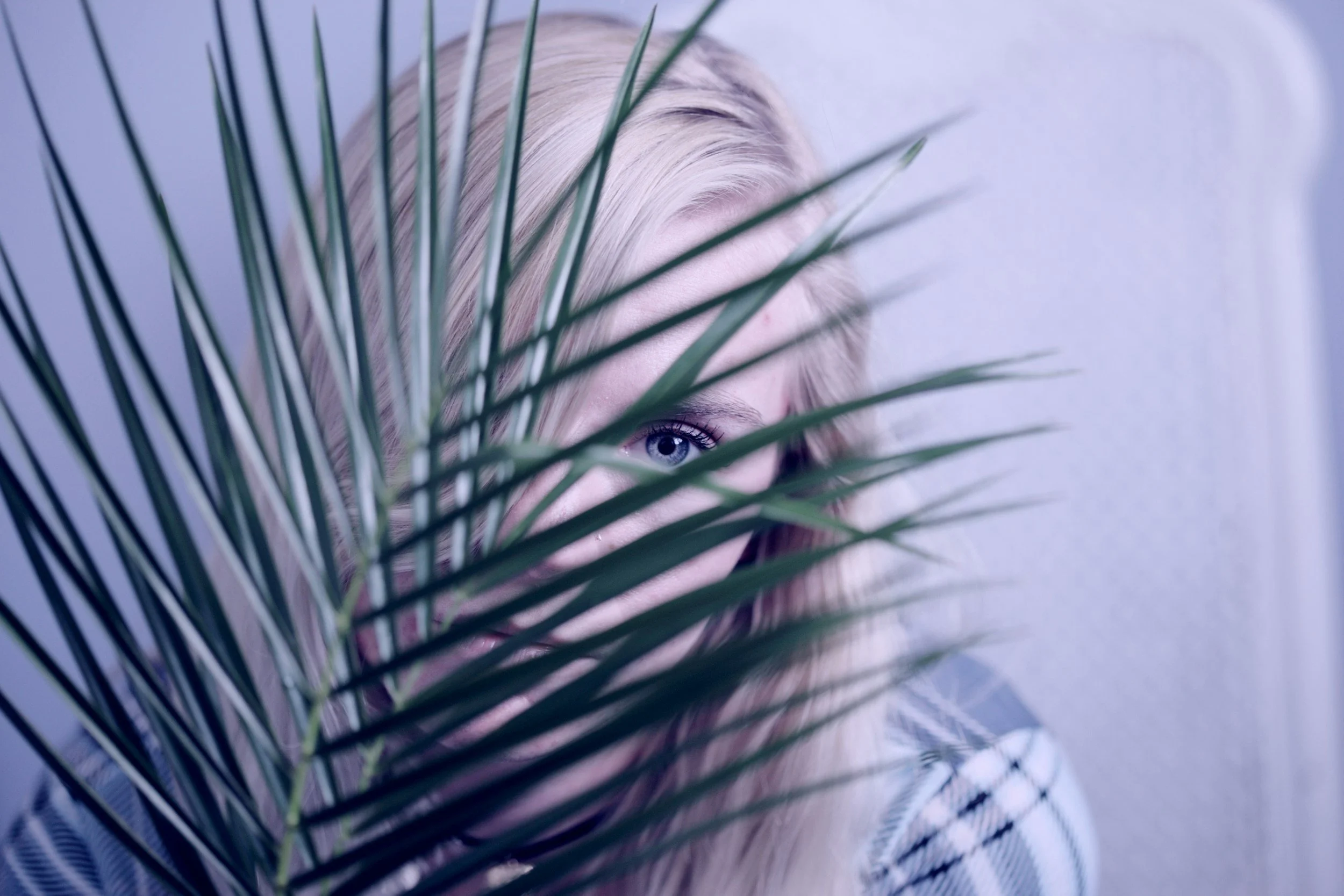 Woman with blonde hair and blue eyes partially hidden behind a large green plant."}