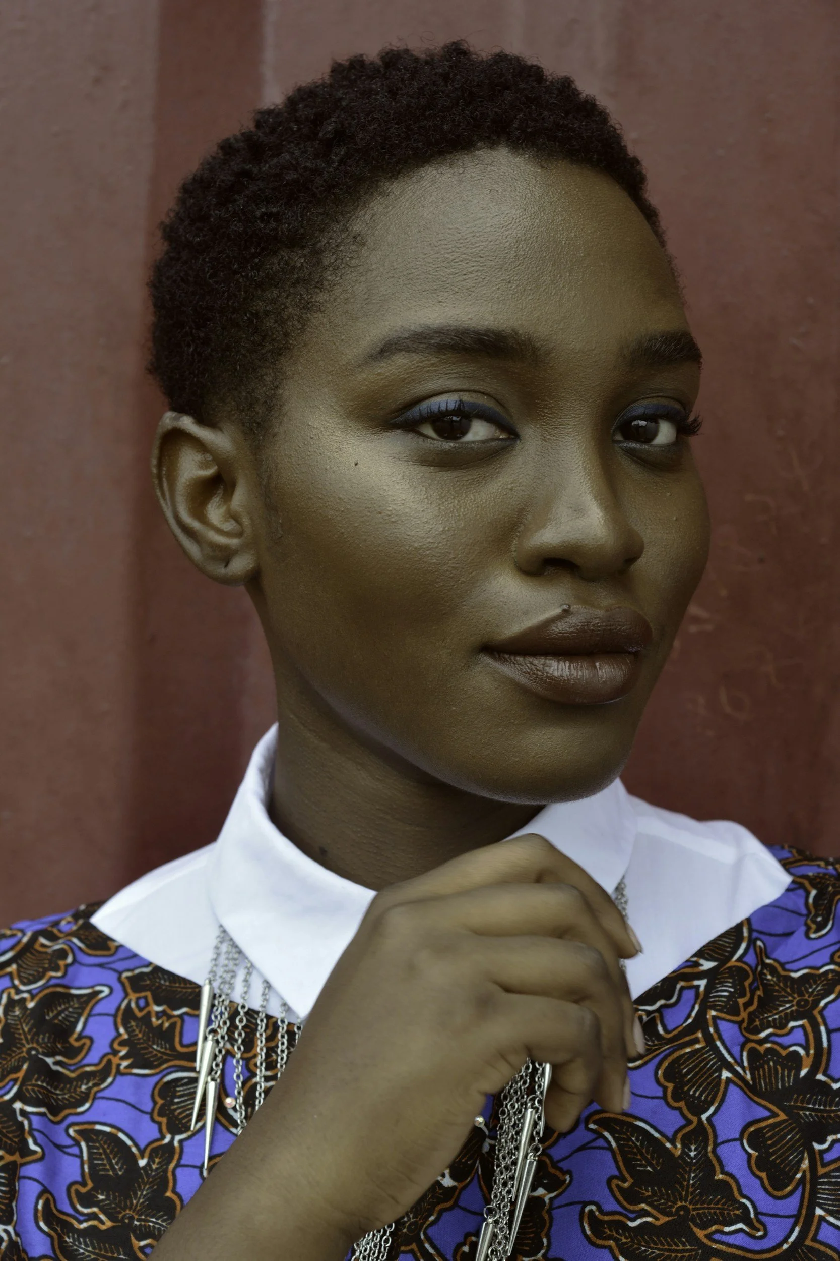 A woman with short natural hair and brown skin, wearing makeup with blue eyeshadow and brown lipstick, dressed in a colorful patterned top with a white collar, standing in front of a reddish-brown wall.