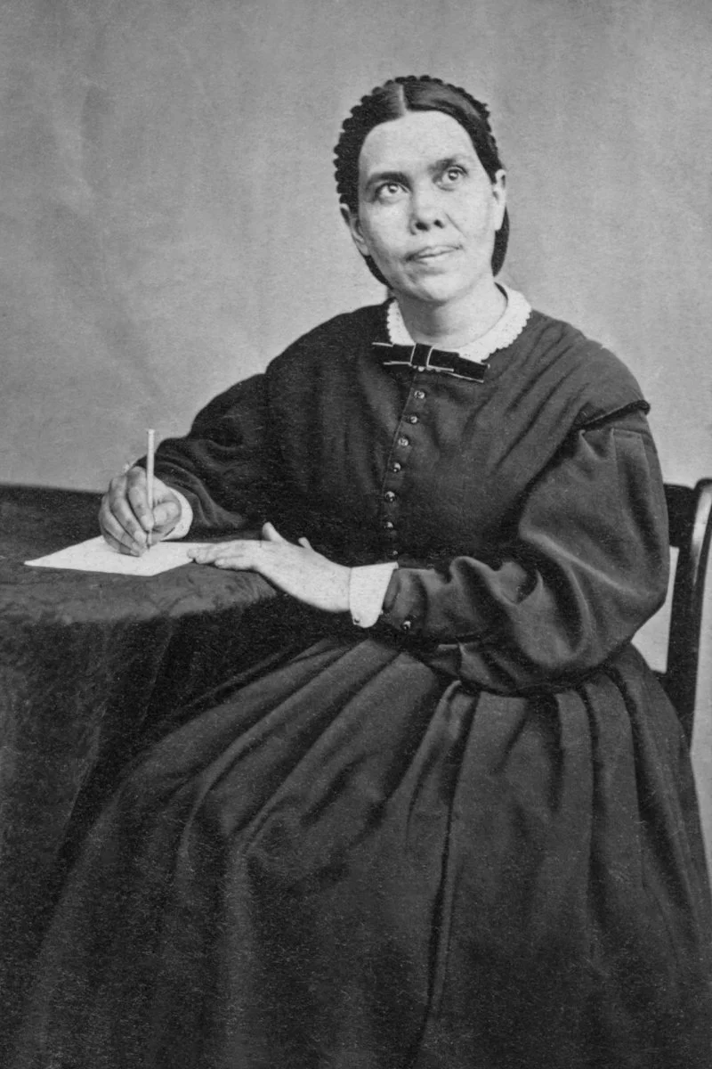 A historical black and white photograph of a woman seated at a table, writing on a sheet of paper with a pen. She is wearing a dark dress with a white collar and appears to be in a thoughtful pose.