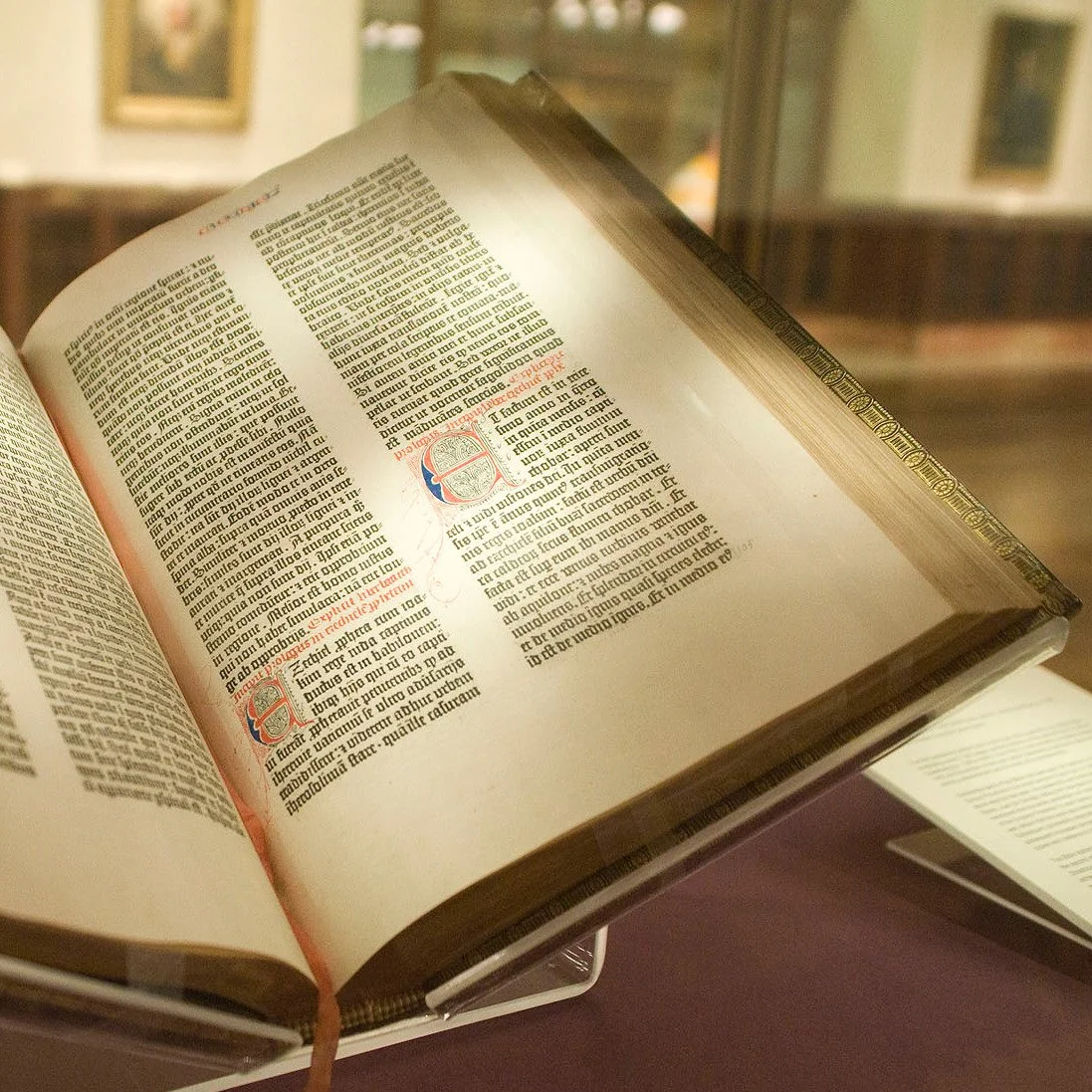 Open illuminated antique book on display in a museum or library with blurred framed pictures in the background.