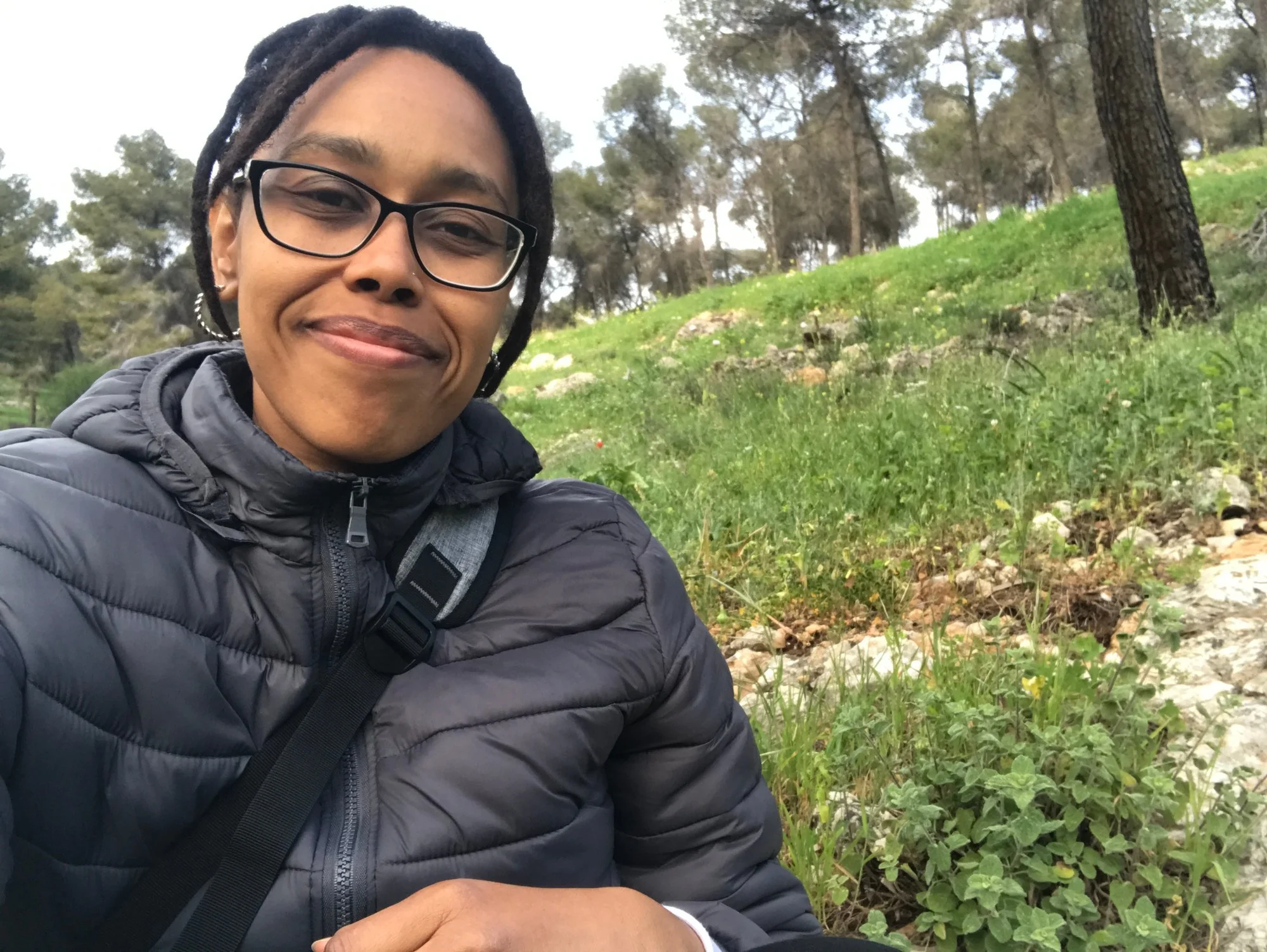 Person smiling outdoors in a wooded area wearing glasses and a puffer jacket.