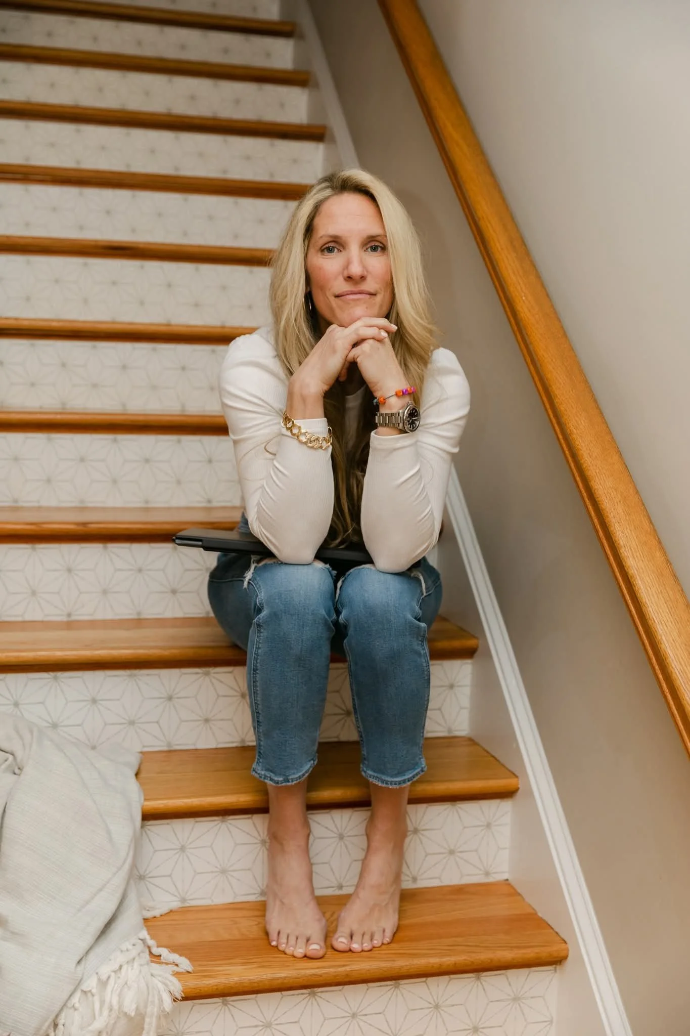 A woman with blonde hair sitting barefoot on a staircase, resting her chin on her hands, wearing a white long-sleeve shirt and ripped jeans.