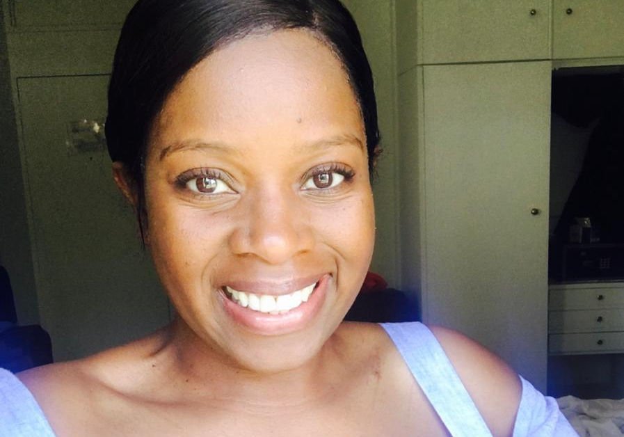 A smiling woman with short dark hair and light skin, wearing a light-colored tank top indoors, with a background of green cabinets and furniture.