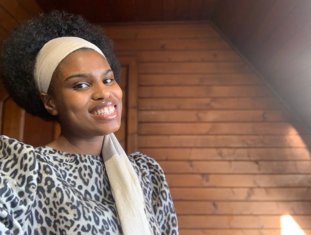 Smiling woman with dark curly hair, wearing a beige headband and leopard print top, in a wooden room with angled ceiling.