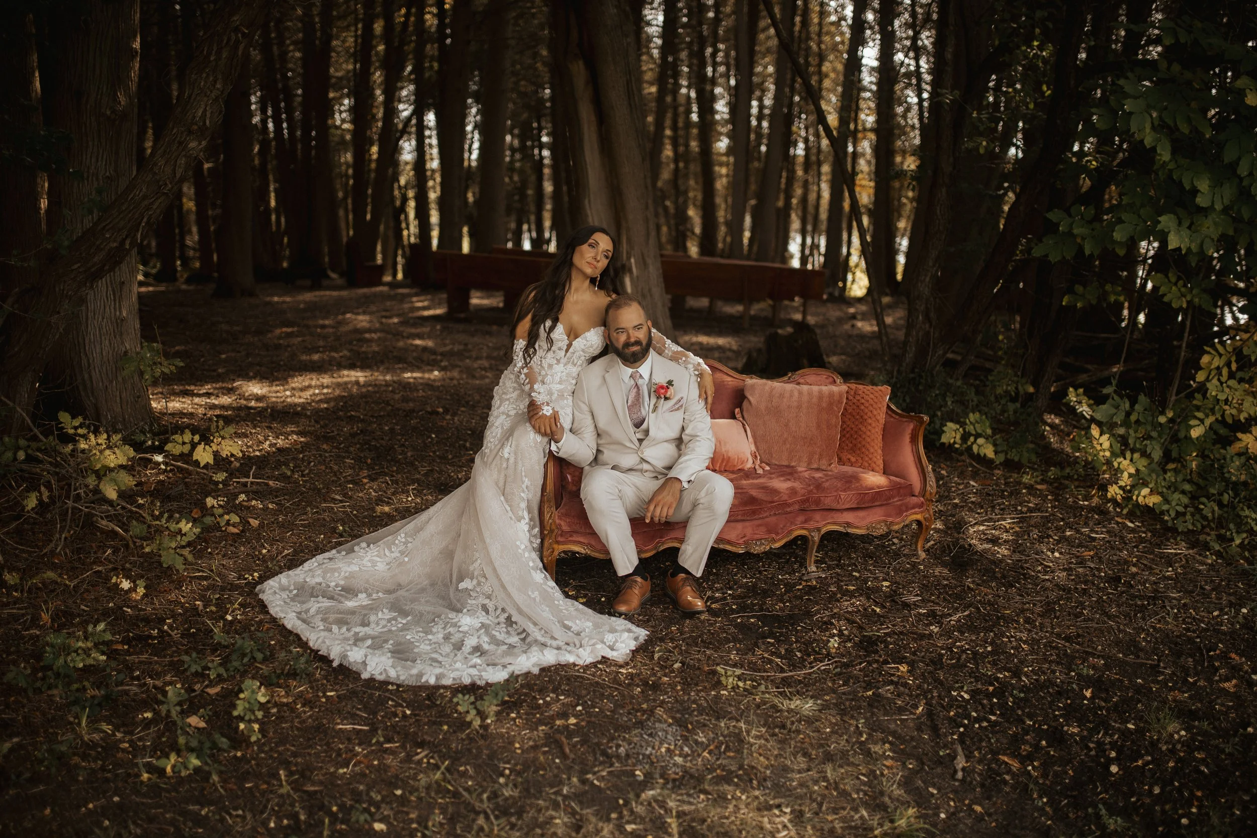A bride and groom sit on a vintage pink sofa in a wooded outdoor setting, with the bride standing beside the groom and her arm around his shoulder. The bride wears a lace wedding gown with long sleeves, and the groom wears a light-colored suit with a
