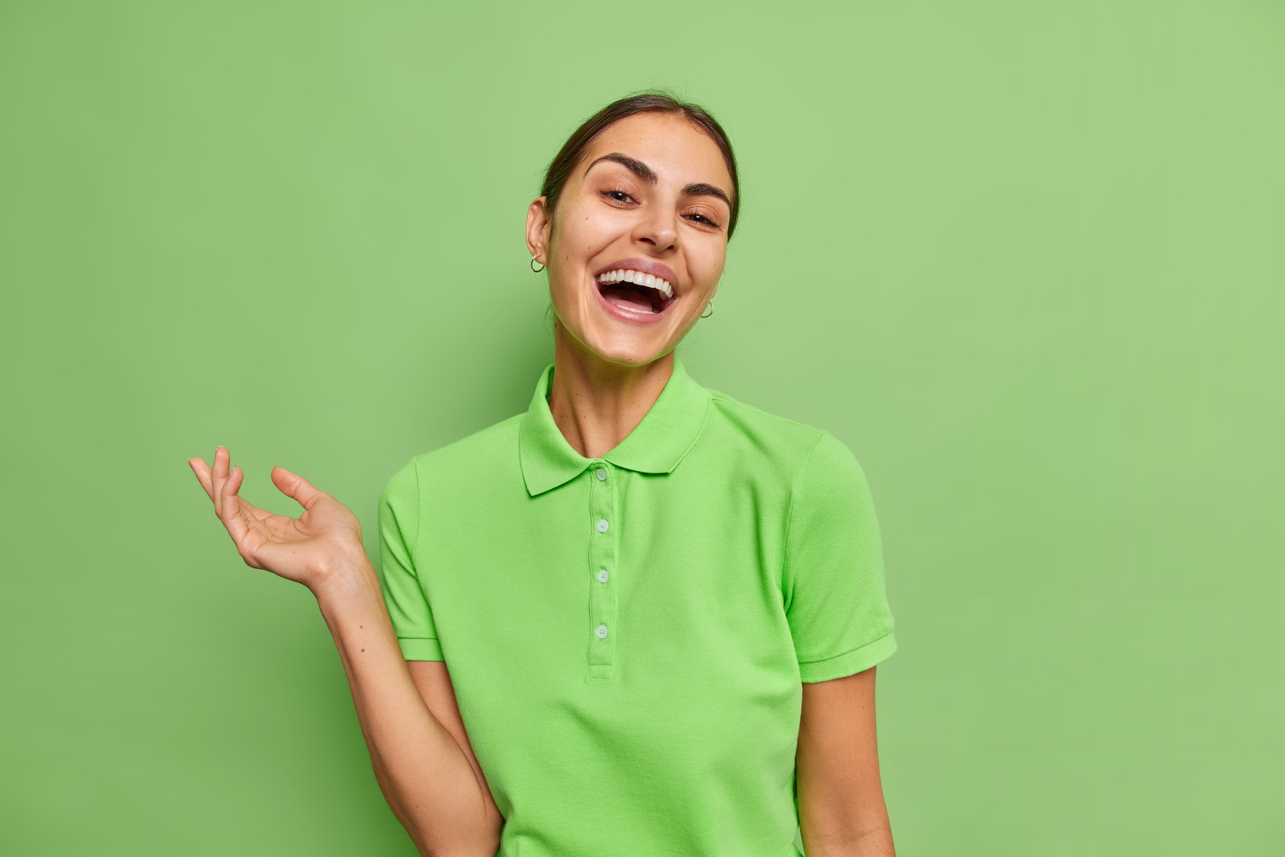 waist-up-shot-happy-brunette-woman-dressed-casual-t-shirt-smiles-sincerely-keeps-arm-raised-laughs-funny-anecdote-wears-casual-t-shirt-isolated-green-background-happiness-concept.jpg