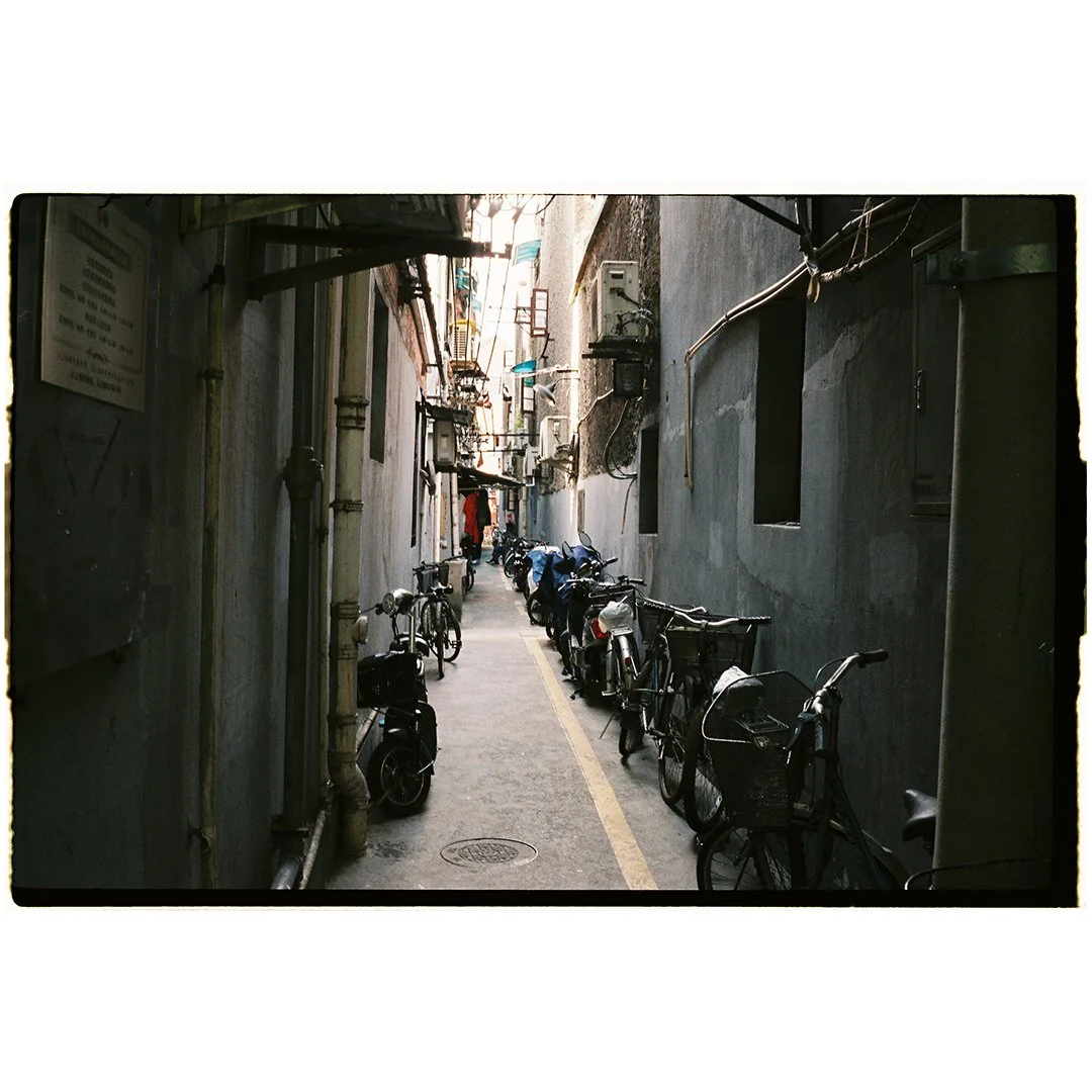 Narrow alleyway with parked bicycles and motorcycles, gray walls, pipes, and utility boxes.